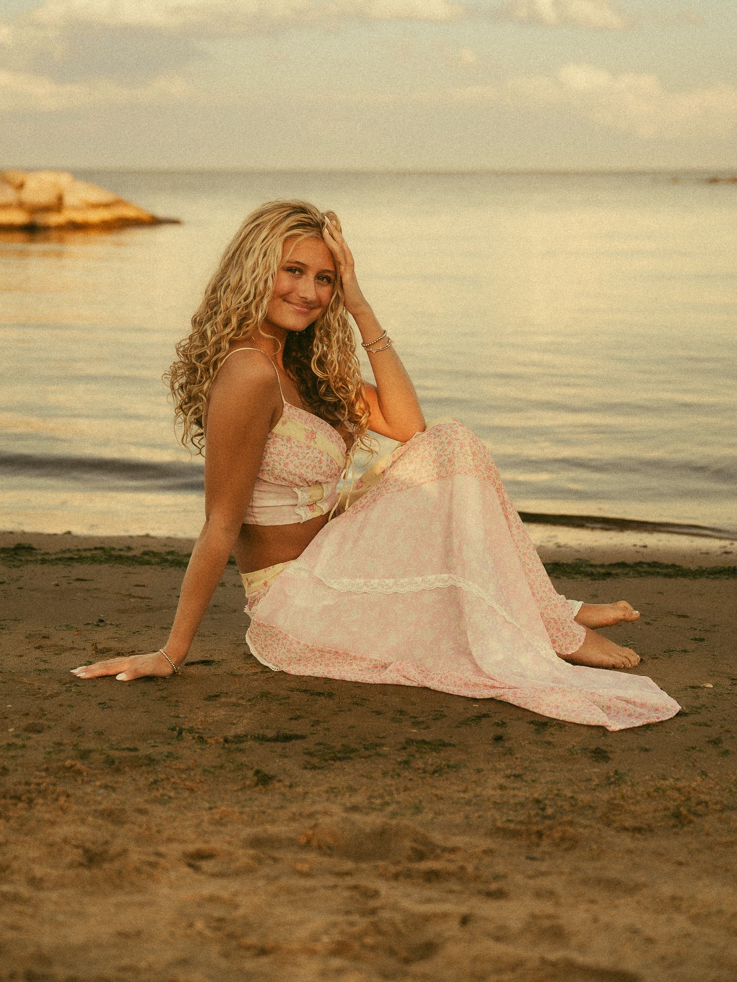 Blonde woman in white crop top and flowy skirt sitting on sandy beach at golden hour sunset.