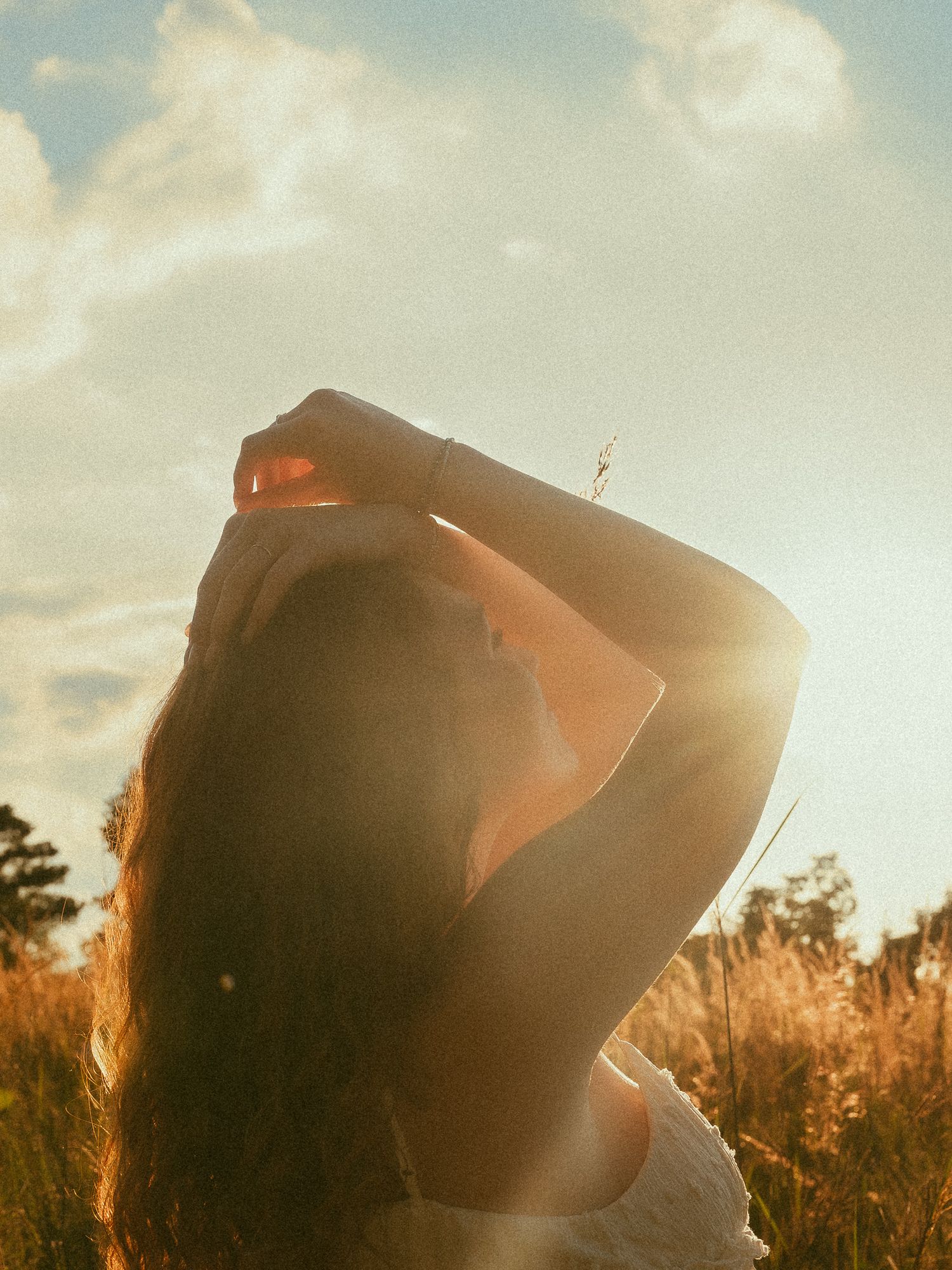 A woman stands in a sunlit field, golden hour light glowing behind her as she raises her hand above her head.
