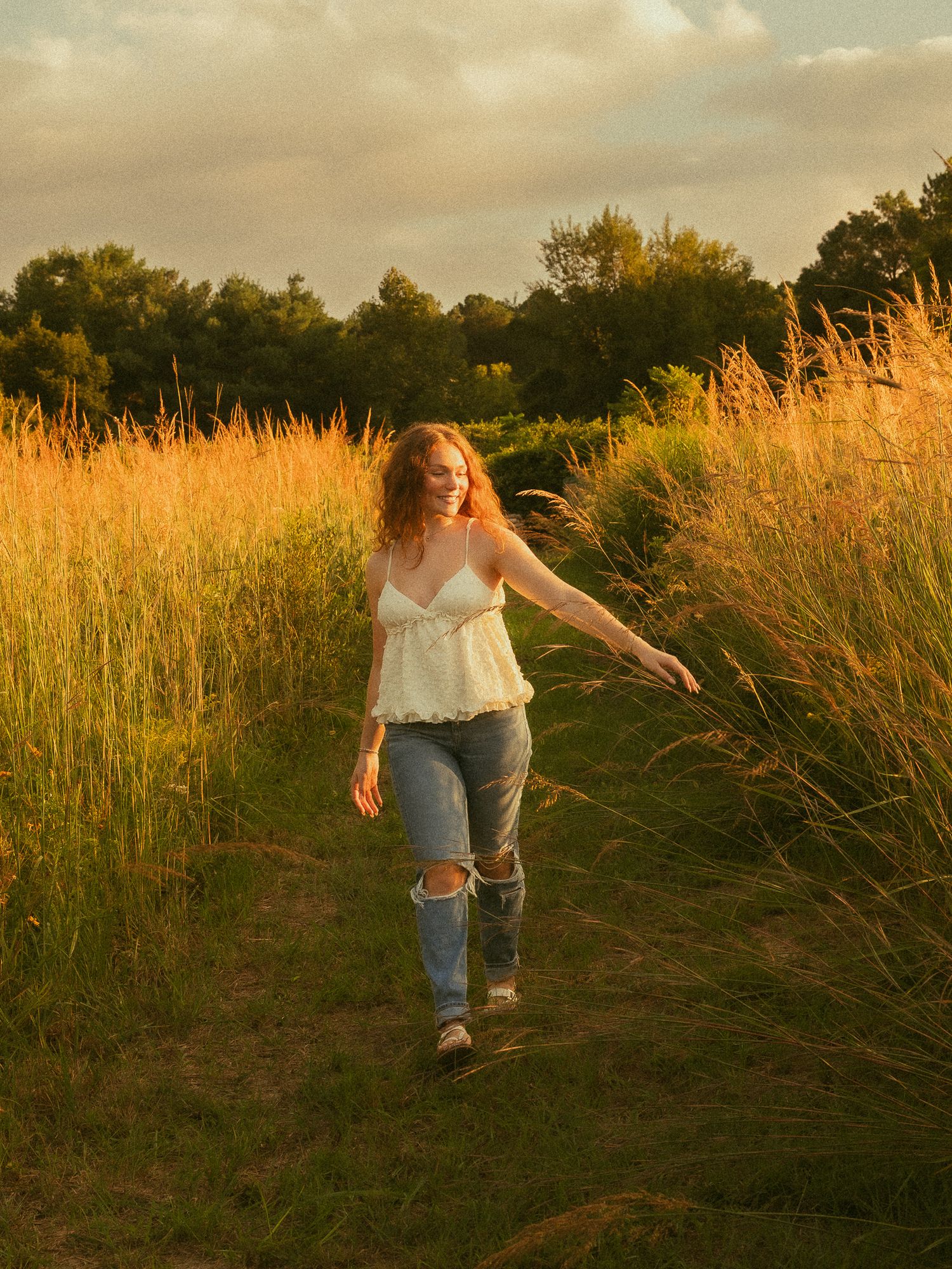 A young woman with red hair walks through a golden sunlit field, wearing a white top and ripped jeans at sunset.