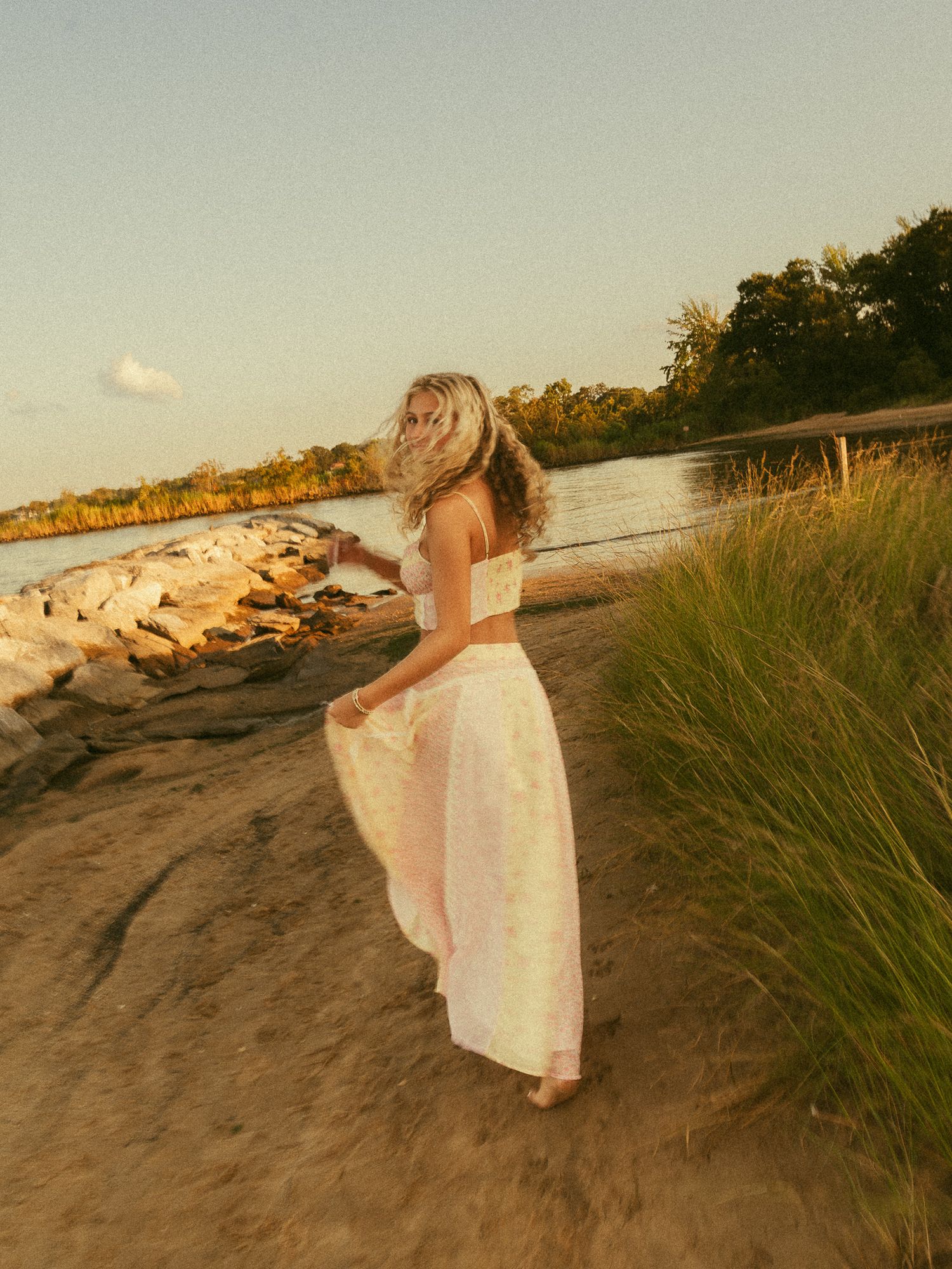A blonde woman in a white crop top and flowy skirt walks along a sandy beach at golden hour.