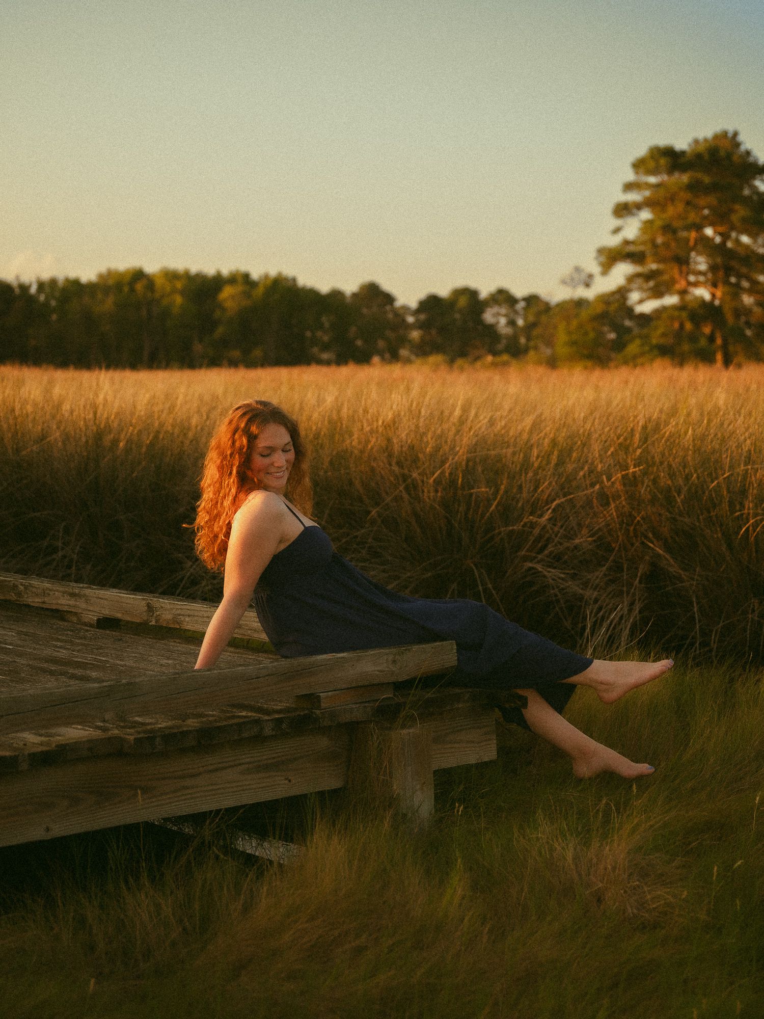 A smiling redhead woman in a dark dress reclines on a wooden boardwalk surrounded by golden marsh grass at sunset.