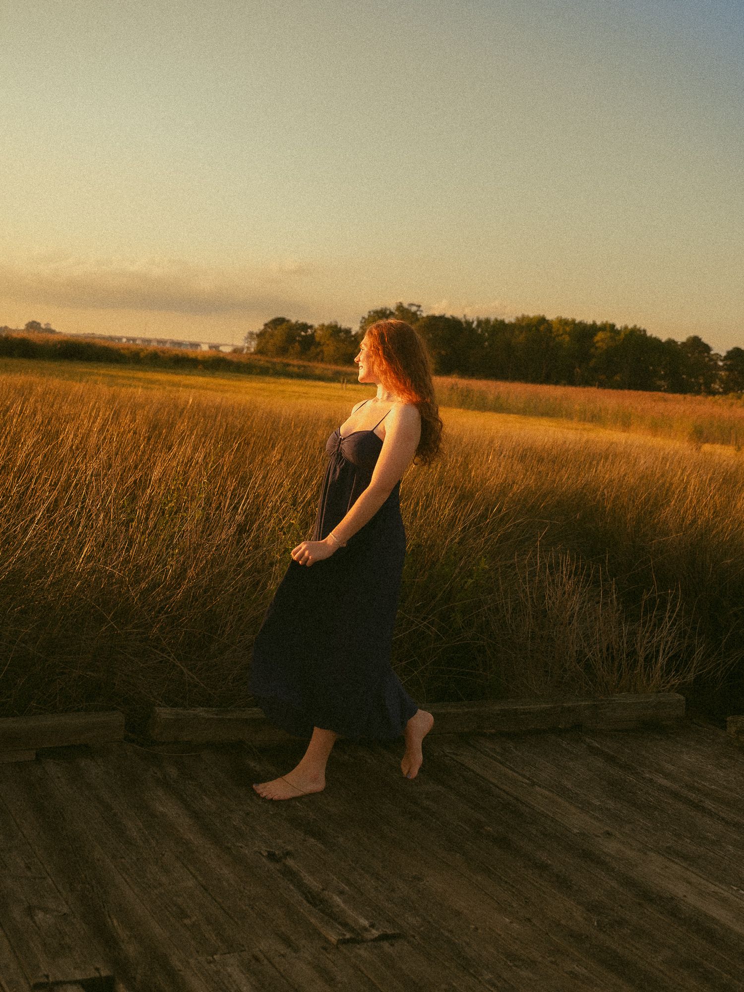 A barefoot woman in a navy dress walks on a wooden dock at golden hour beside tall golden marsh grass.
