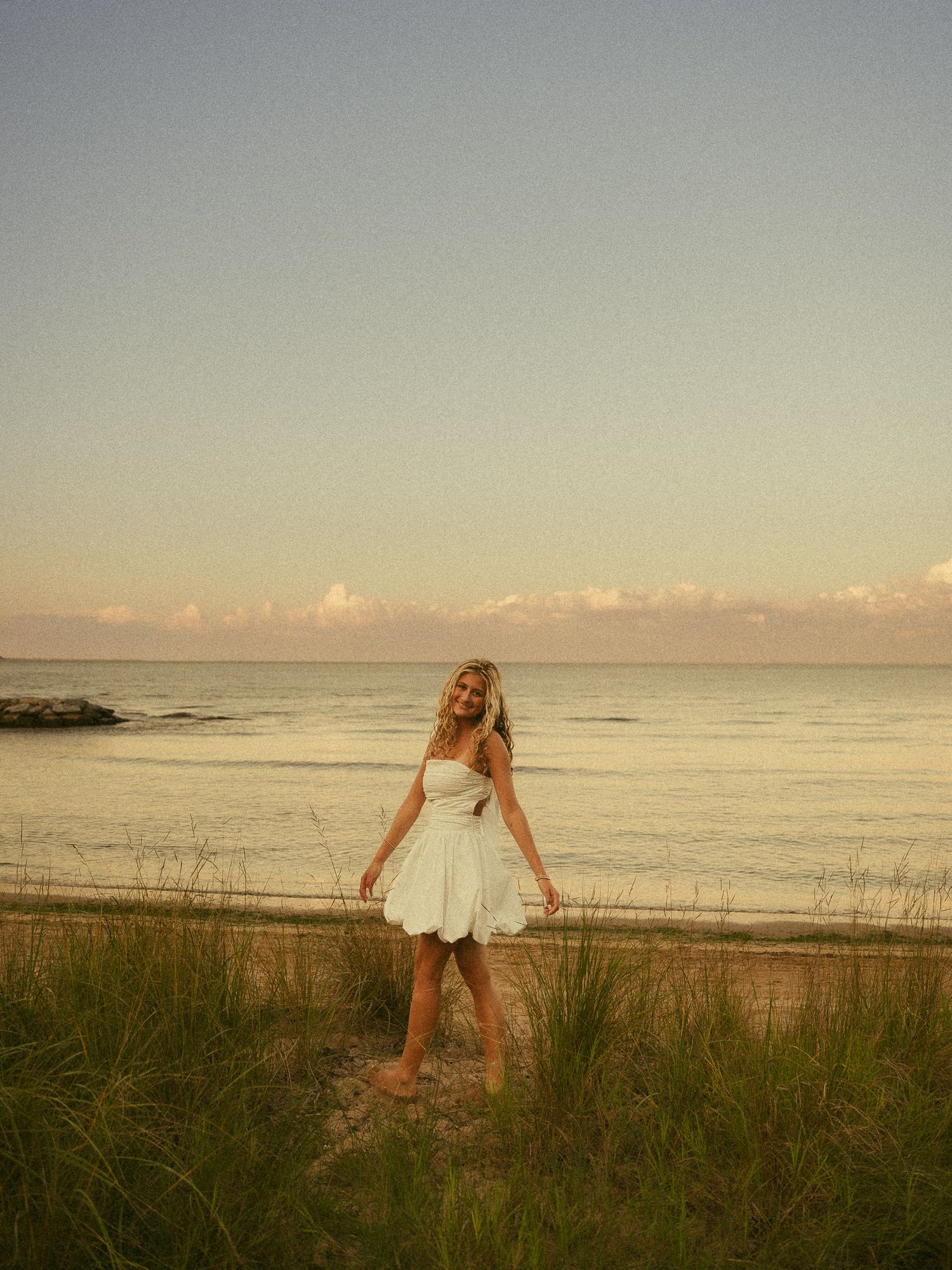 Blonde woman in white mini dress standing on beach path with ocean and pastel sky behind her.