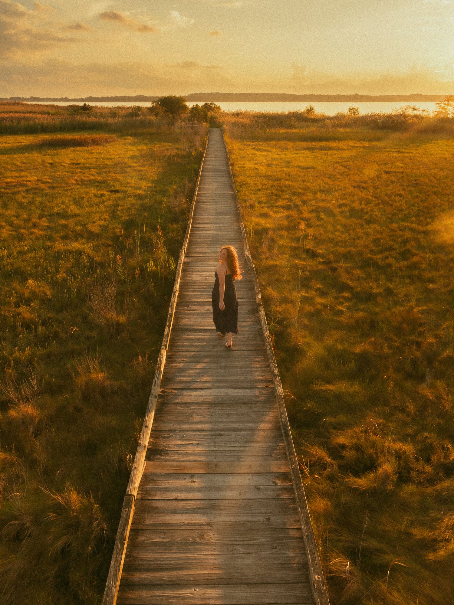 Woman in black dress walking along a wooden boardwalk through golden marsh grass at sunset.