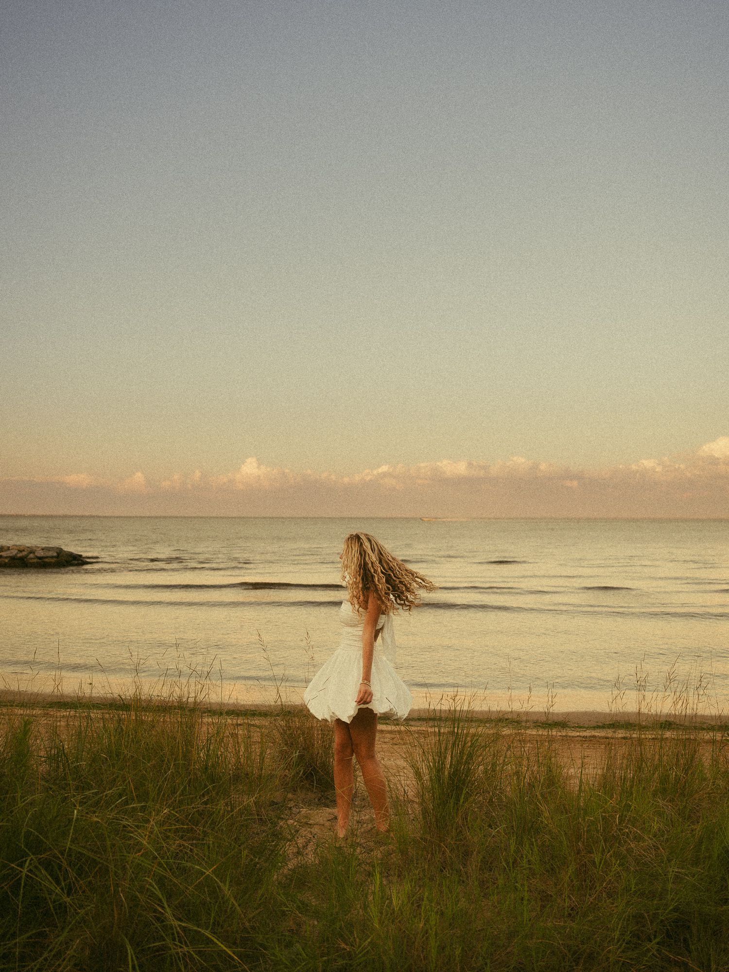 Woman in white dress standing on beach at sunset with hair blowing in the wind.