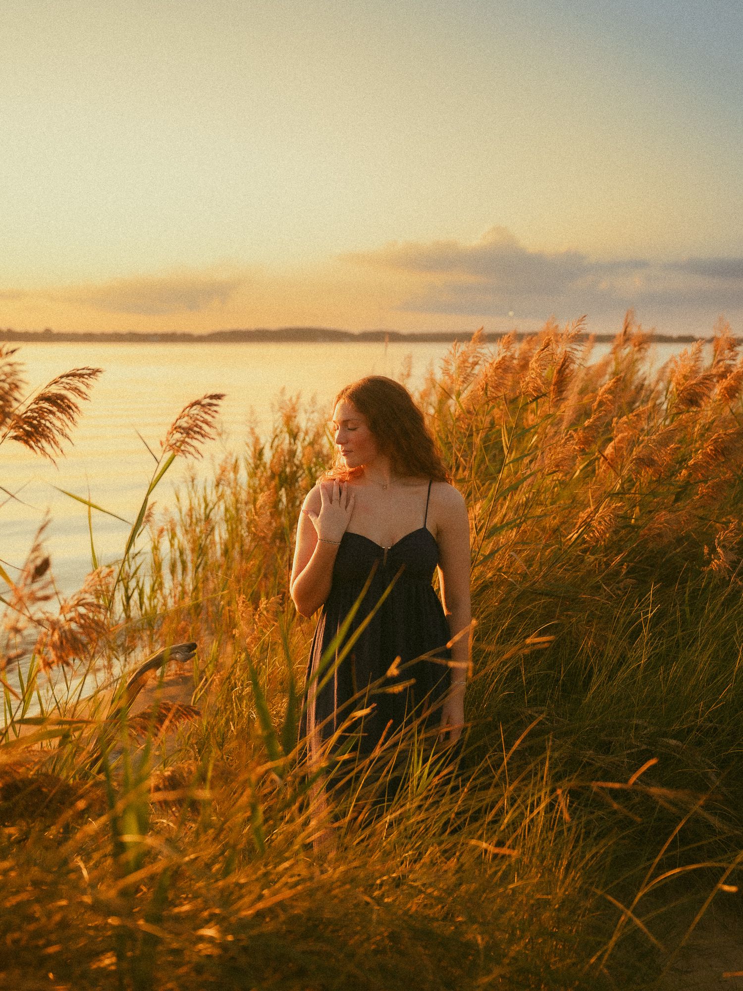Woman in black dress standing among golden grass by a lake at sunset.