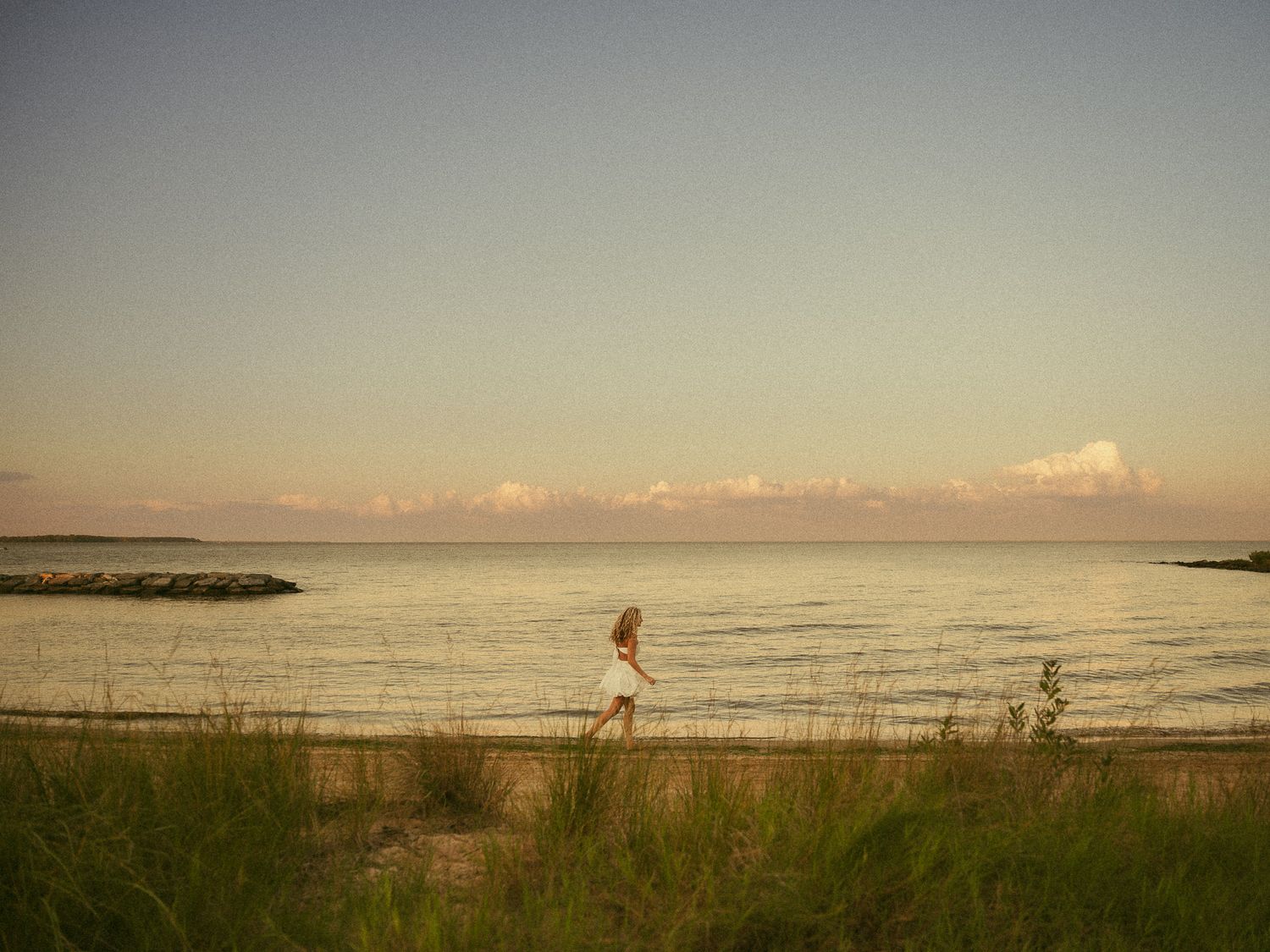 A woman in a white dress walks along a sandy beach path at sunset with calm ocean waters in the background.