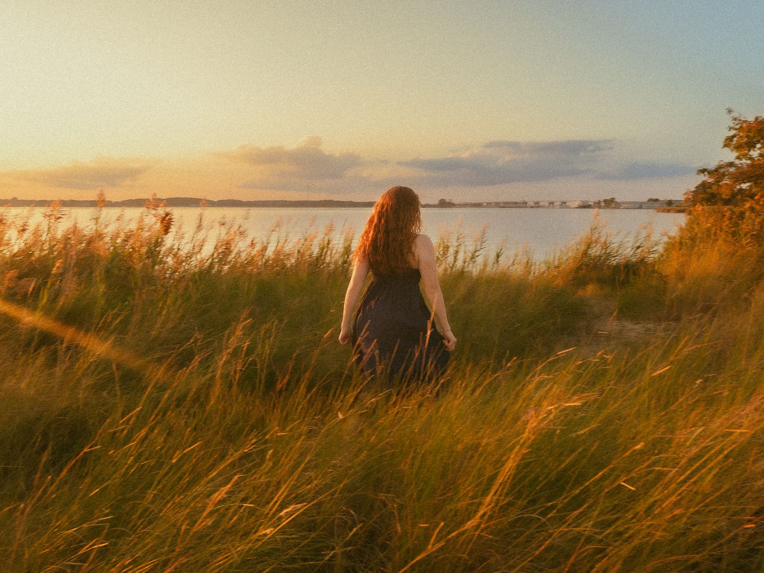 Woman with red hair standing in golden tall grass fields at sunset near a tranquil lake.