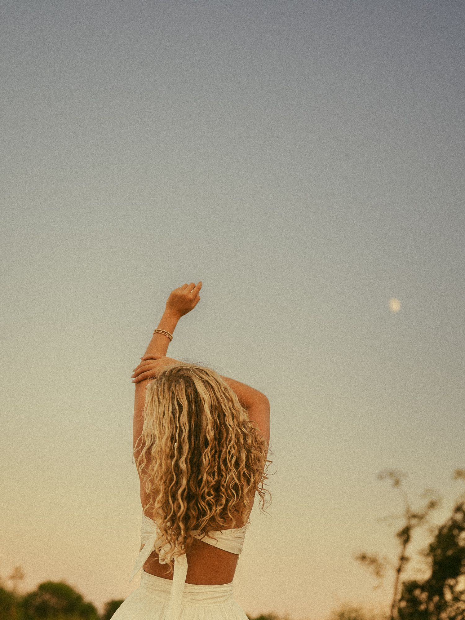 Woman with curly blonde hair pointing at the moon during golden hour sunset outdoors.