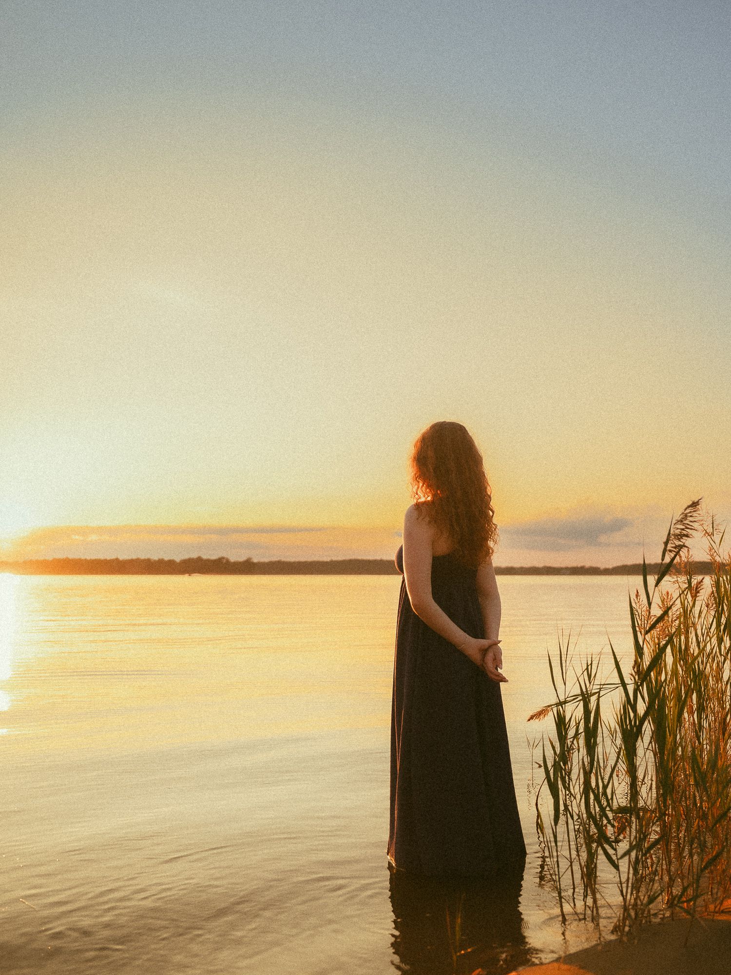 Woman in black dress standing in water at golden sunset by a serene lake with reeds.