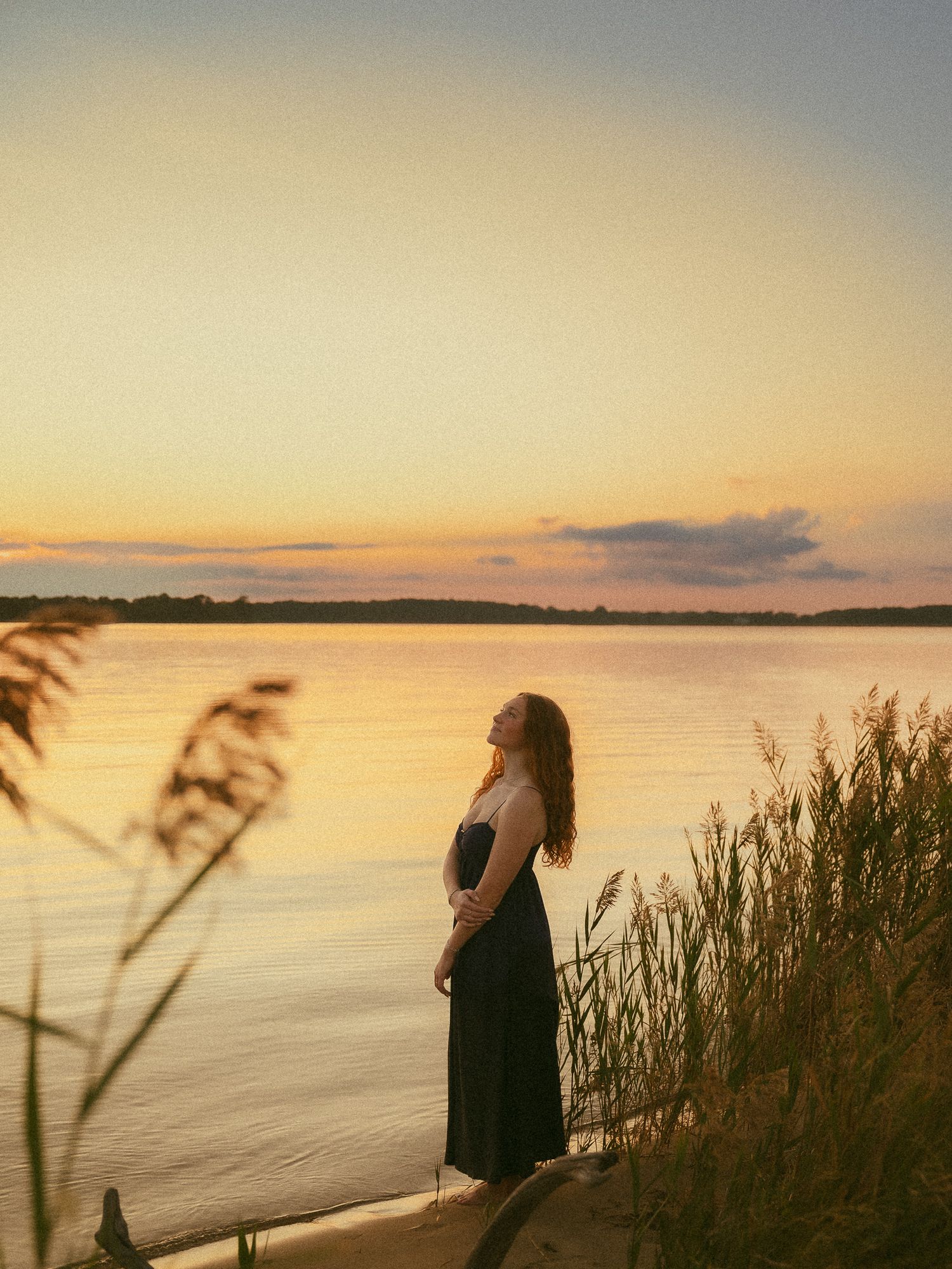 Woman in black dress standing by a serene lake at golden sunset, with tall reeds framing the peaceful waterside scene.