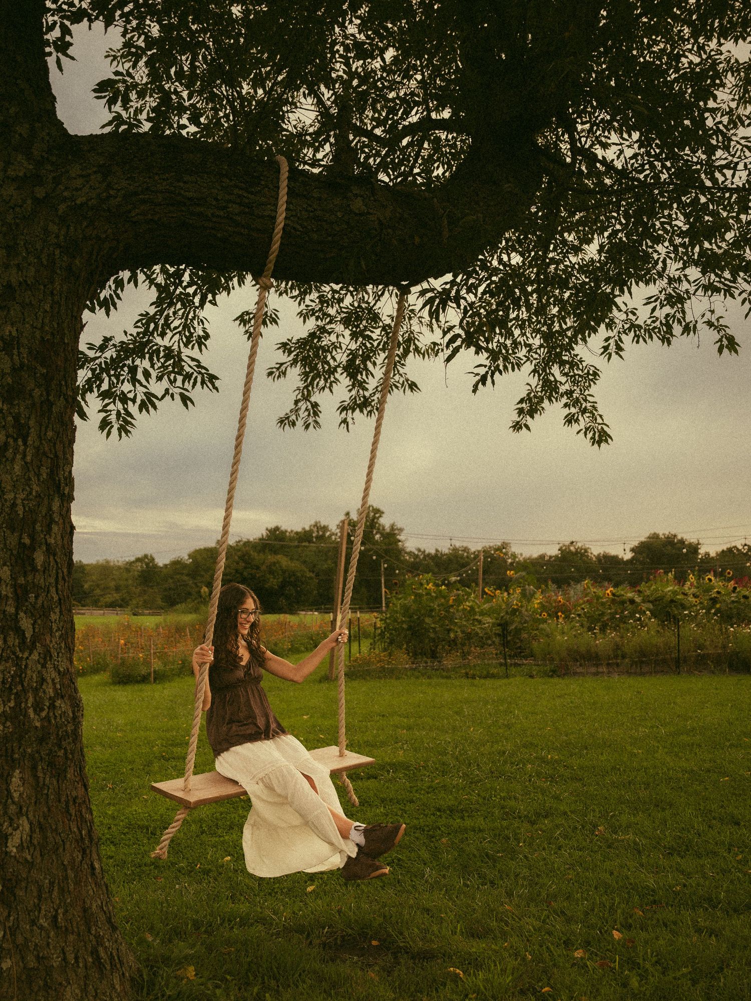 Woman in white skirt sitting on a wooden rope swing hanging from a large tree in a lush green countryside.