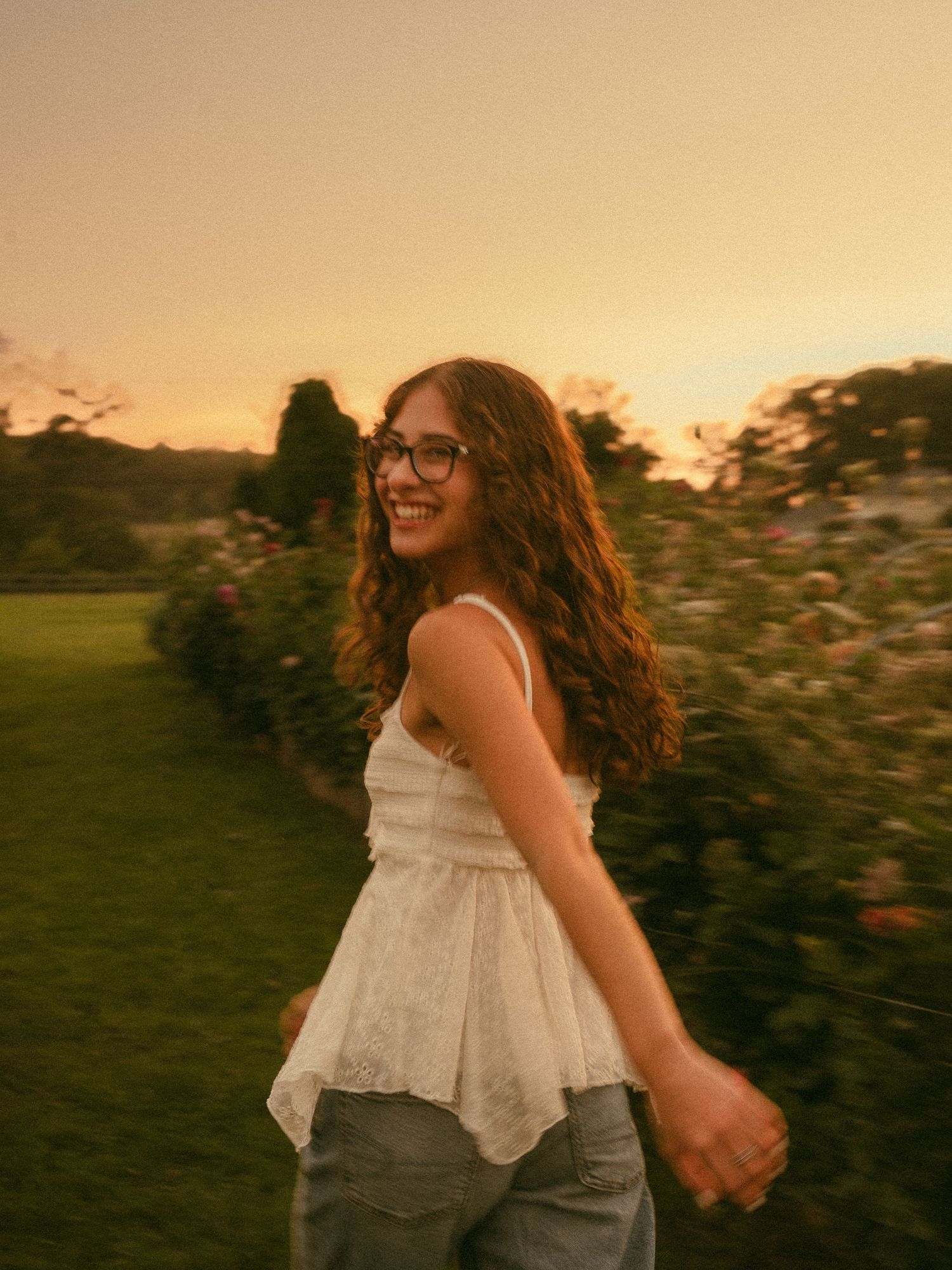 Smiling young woman with curly hair and glasses wearing a white top, spinning outdoors at golden hour sunset.