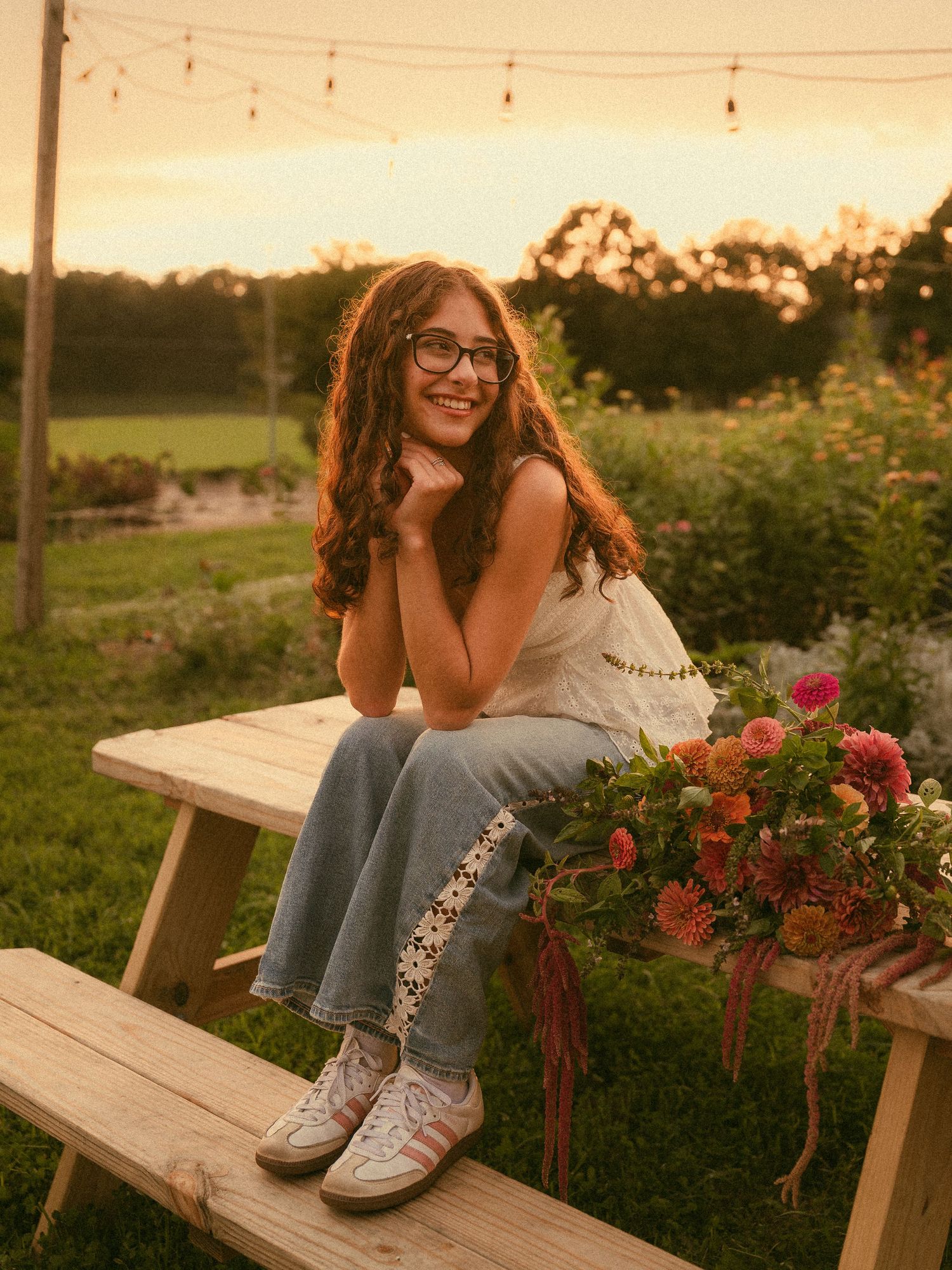 A smiling young woman with glasses sits on a wooden picnic table at sunset, surrounded by colorful flowers in a garden.