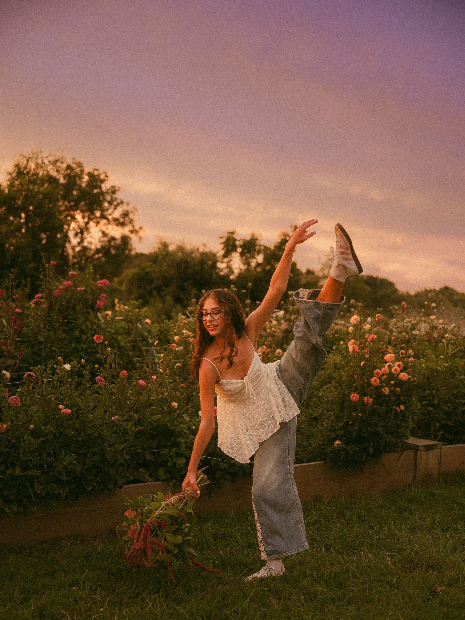 A woman dances joyfully in a flower garden at sunset, holding blooms, wearing white top, jeans, and sneakers.