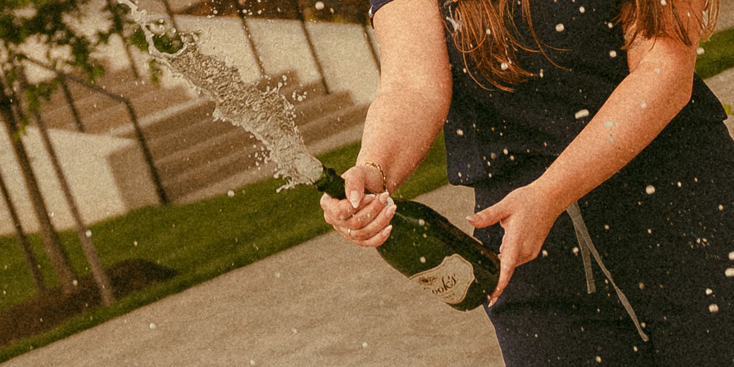 Person spraying champagne outdoors, celebrating with bubbly liquid splashing in the air near a garden.