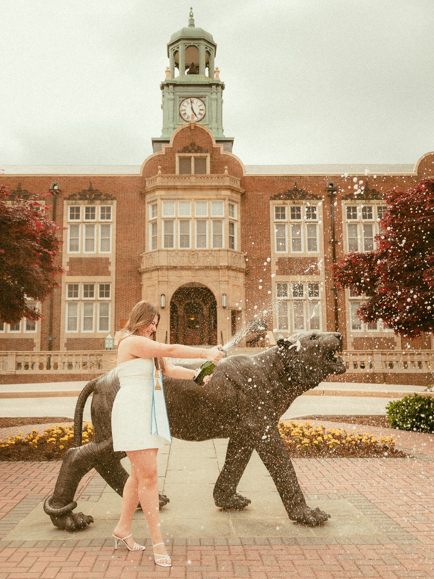 Woman in blue dress popping champagne beside a black panther statue in front of a brick university building.