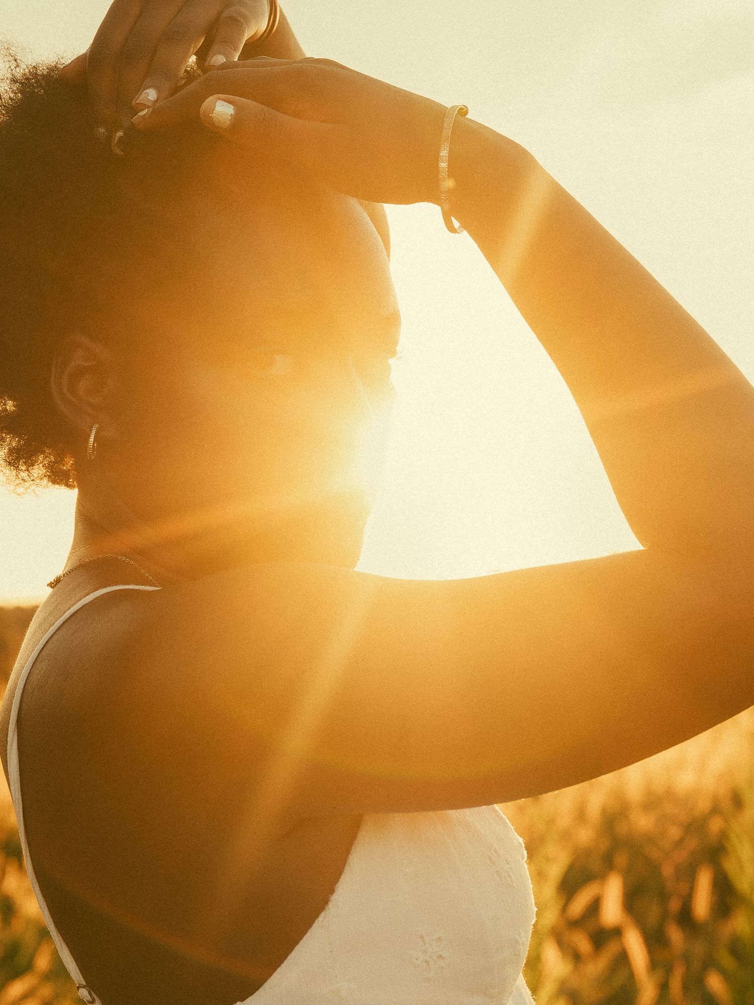 A Black woman stands in a golden sunlit field, hands raised to her hair, backlit by a warm glowing sunset.