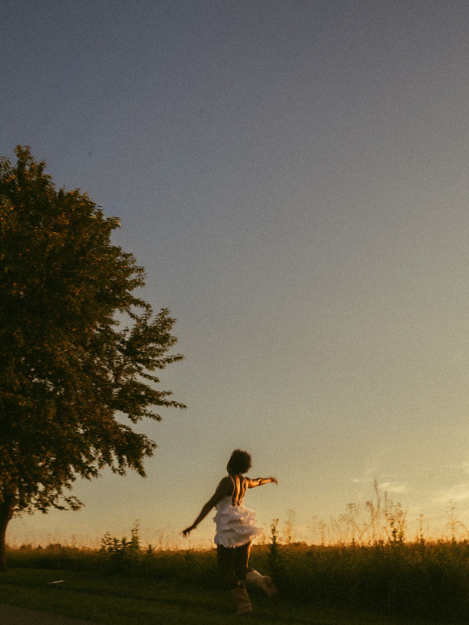 A child in a white dress twirls joyfully in a golden sunset field beside a large silhouetted tree.