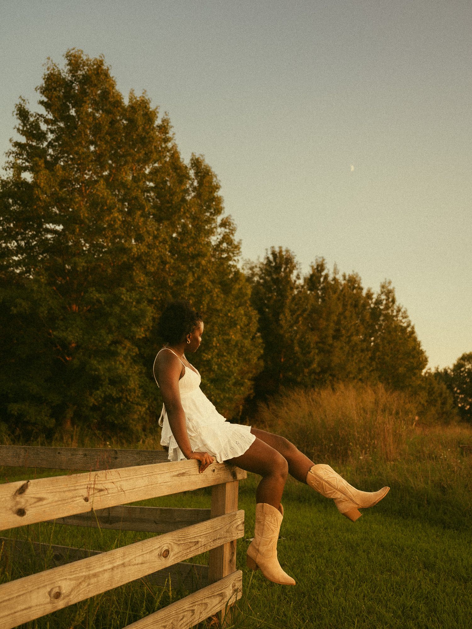 Woman in white dress and cowboy boots sitting on wooden fence at golden hour with trees and crescent moon.