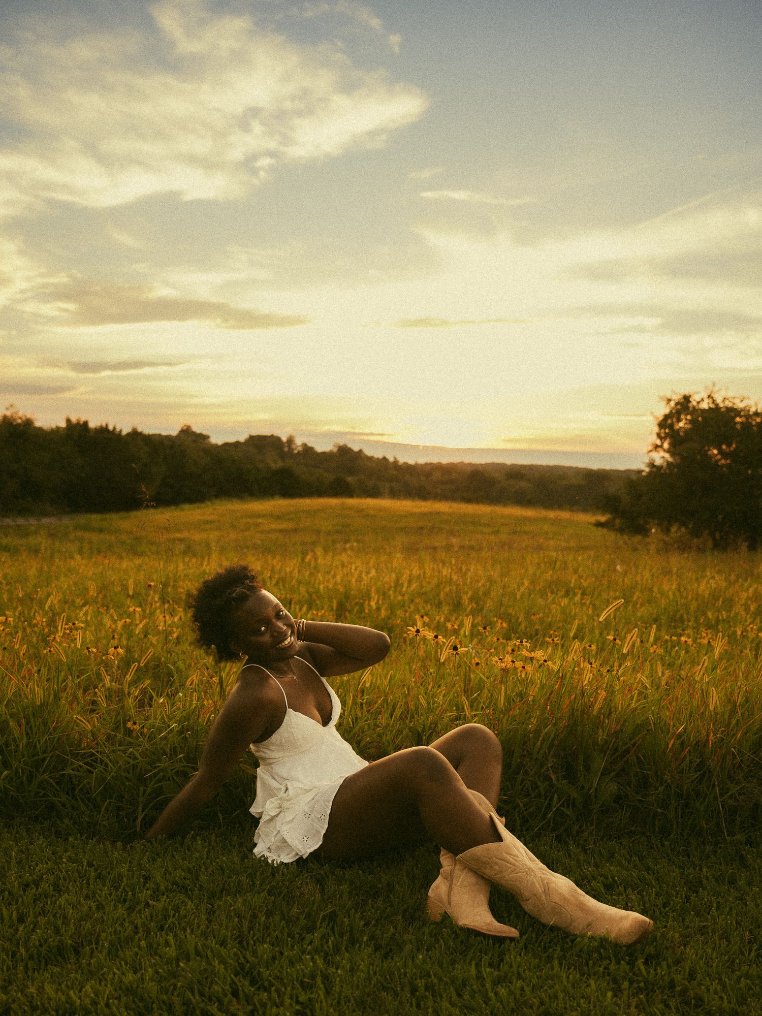 A Black woman in a white dress and cowboy boots sits in a golden meadow at sunset.