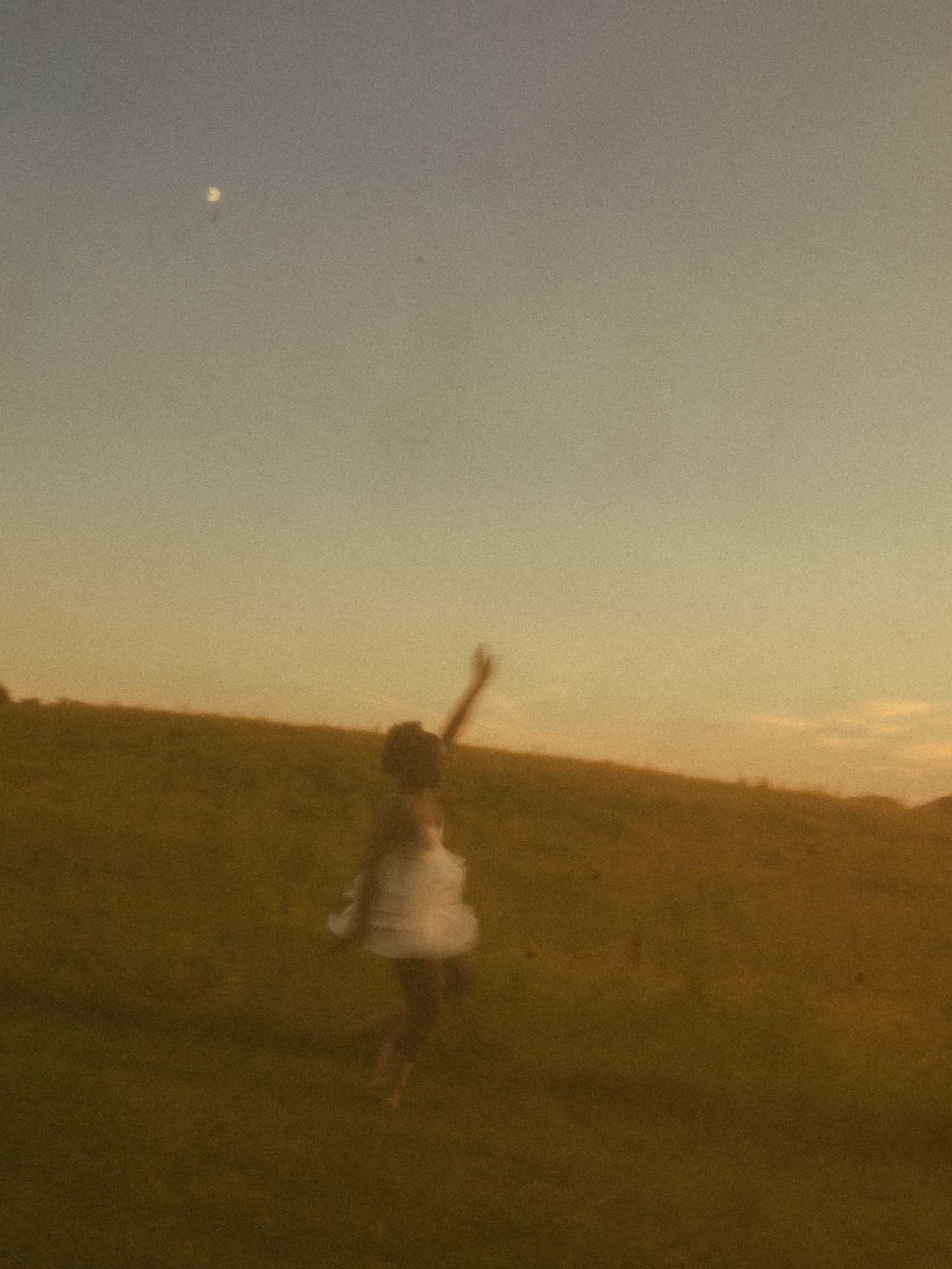 A child in a white dress reaches up toward the moon at dusk in an open field bathed in golden evening light.