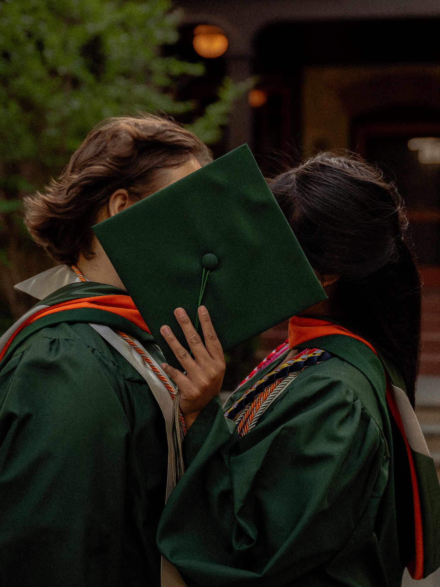 Two graduates in green robes share a kiss hidden behind a graduation cap.
