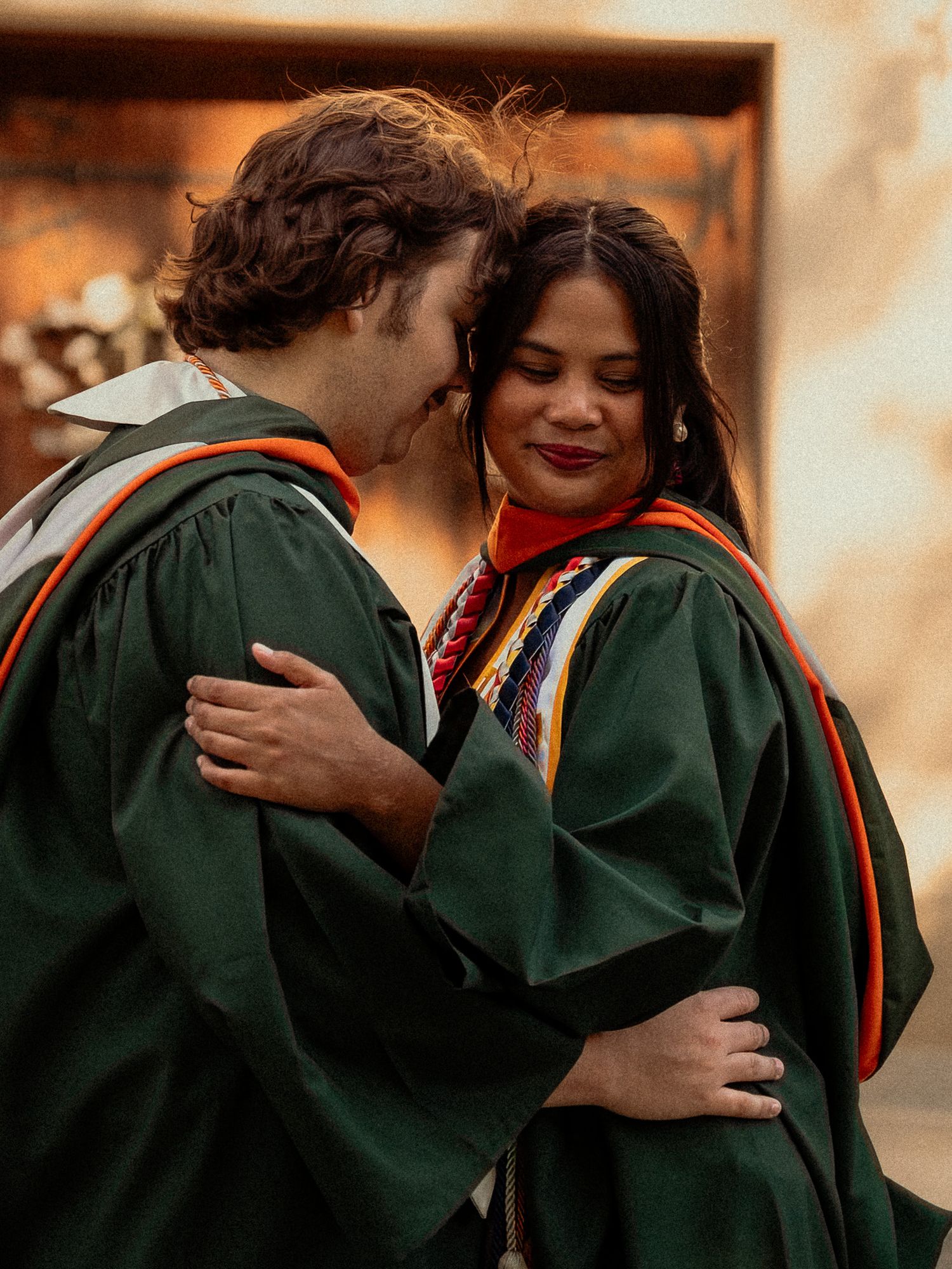 Two smiling graduates in green robes share a warm embrace at graduation.