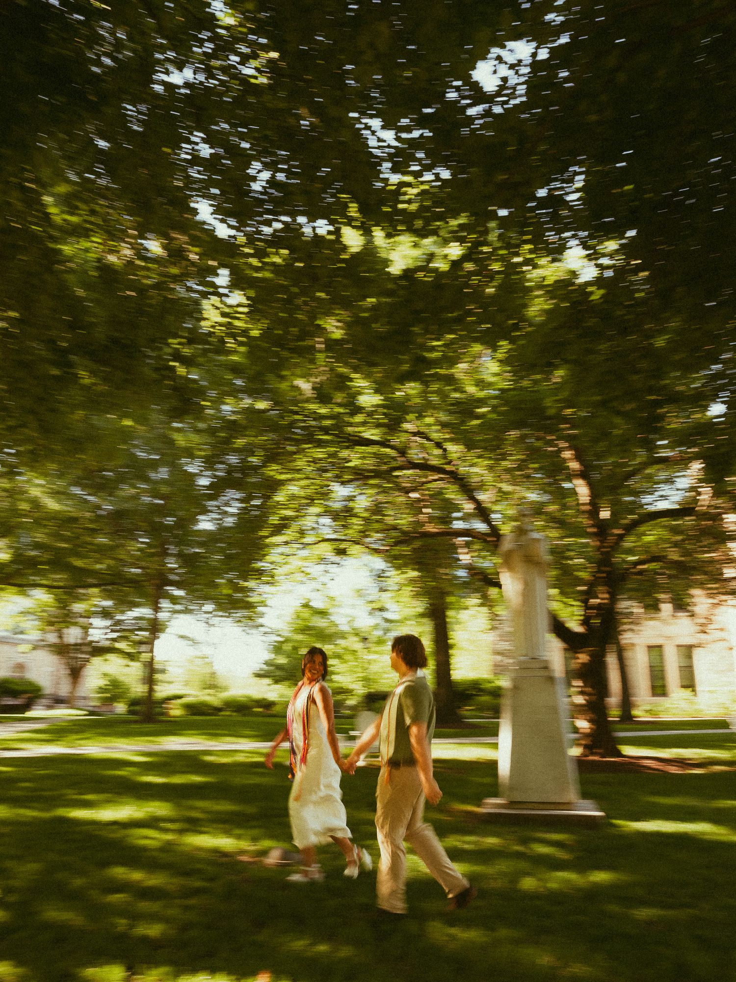 Couple walking hand in hand through a sunlit park with lush green trees and manicured lawn.