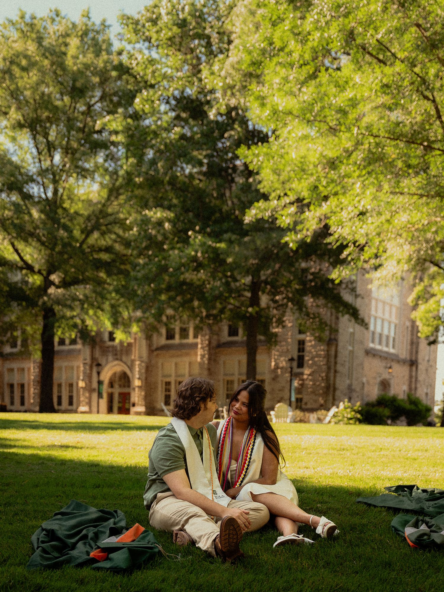 Two graduates in regalia sit on green campus lawn, conversing beneath large trees near a stone university building.