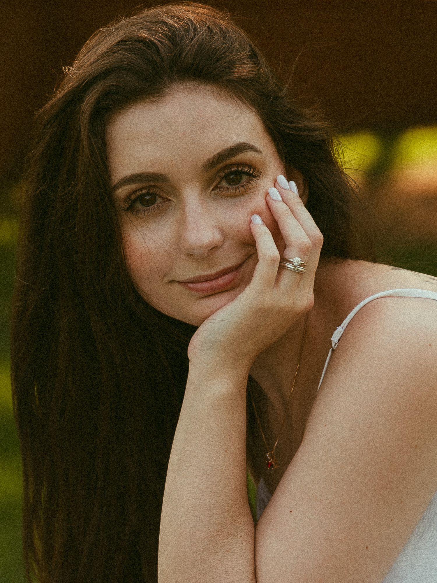 Young brunette woman resting her cheek on her hand, wearing a ring, with a soft smile outdoors.