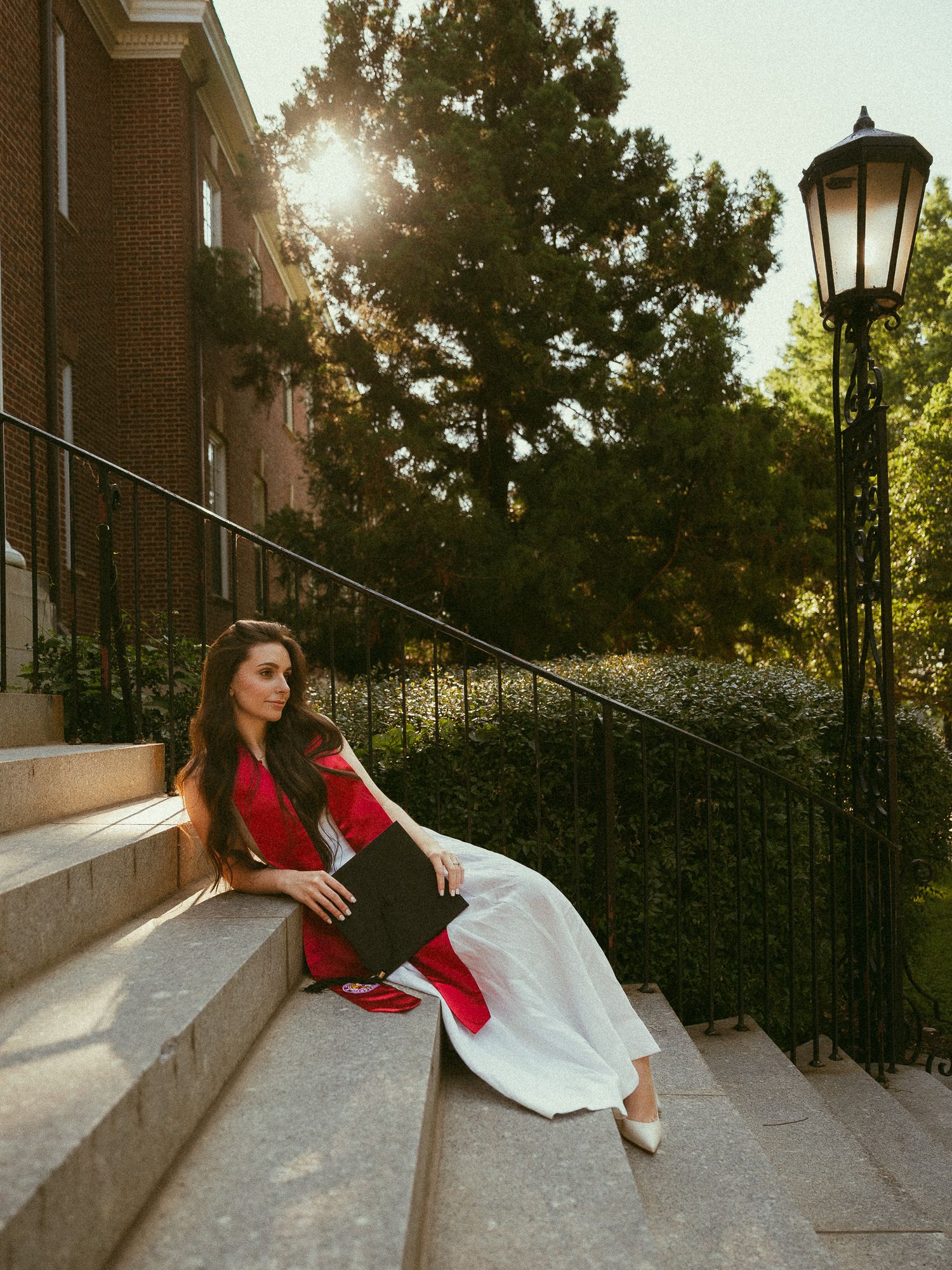 Woman in white dress and red sash sits gracefully on stone steps beside a lamp post at golden hour.