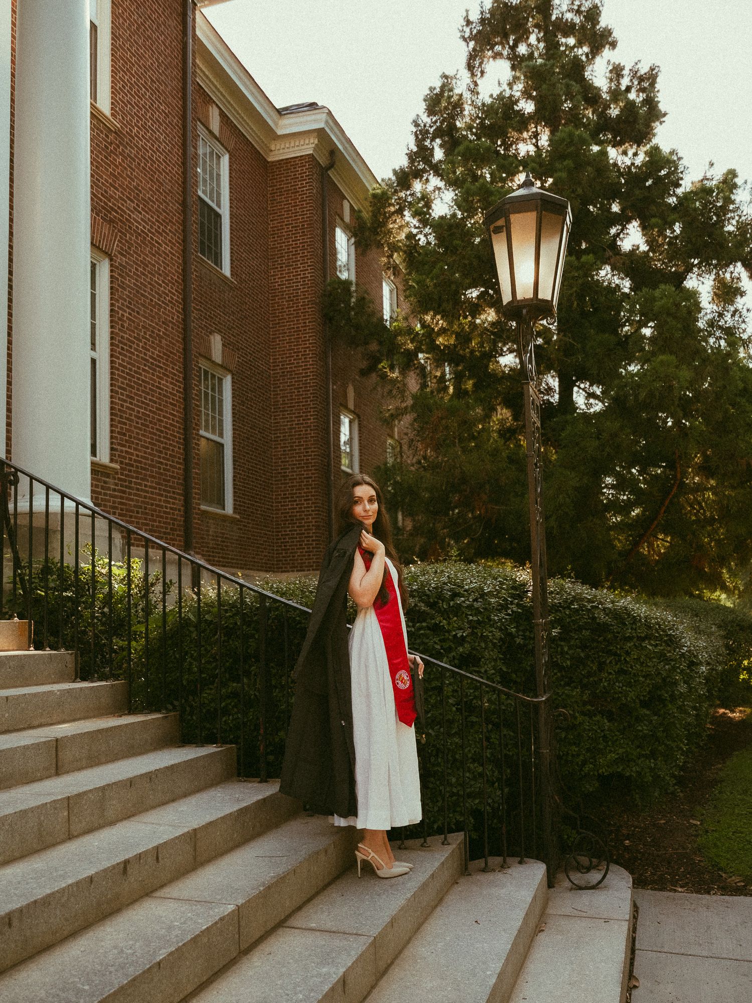 Graduate in black cap and gown with red sash stands on stone steps beside a vintage lamp post.