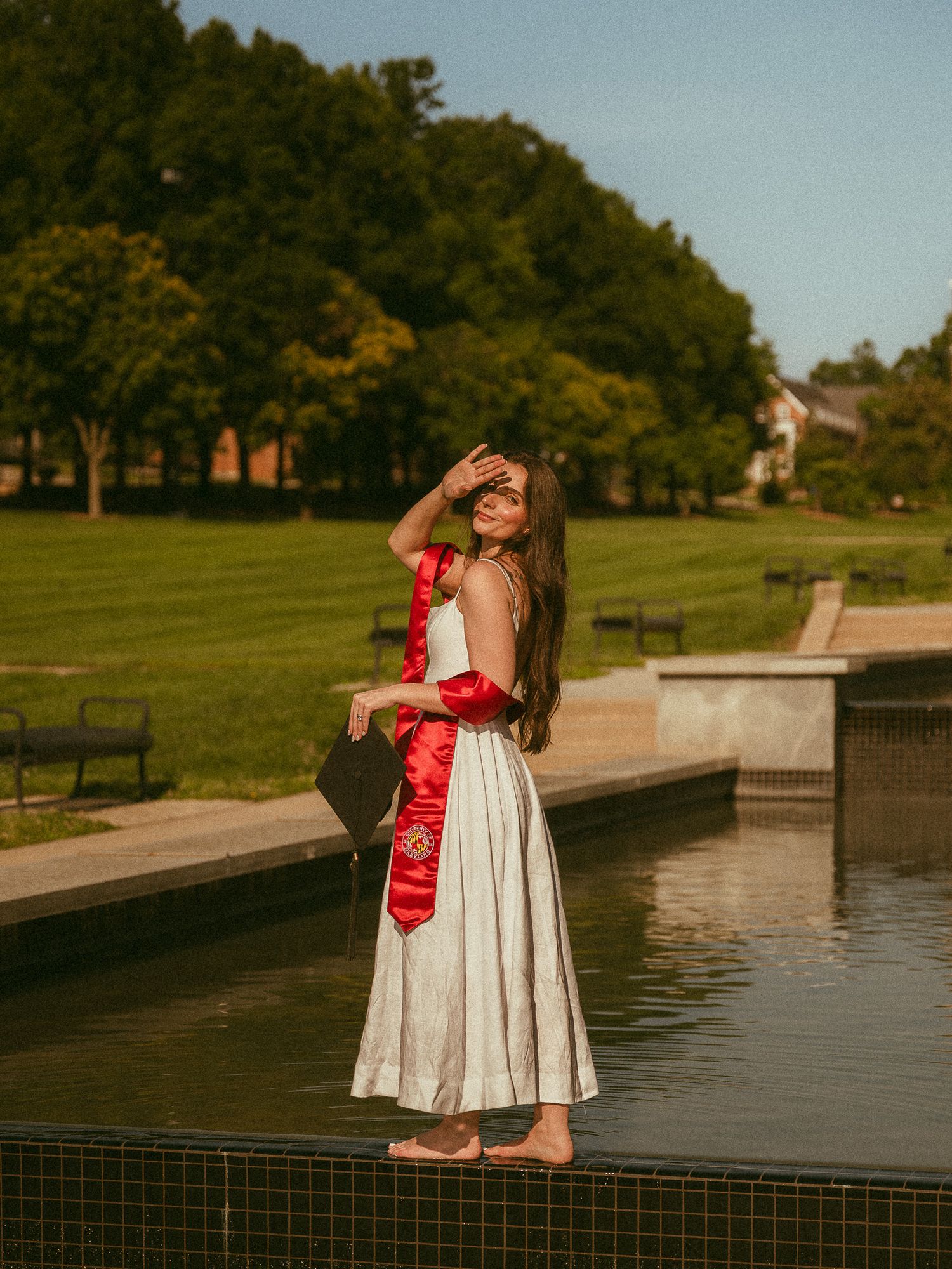 Graduate in white dress holds cap and red sash beside a reflective pool on a sunny campus.