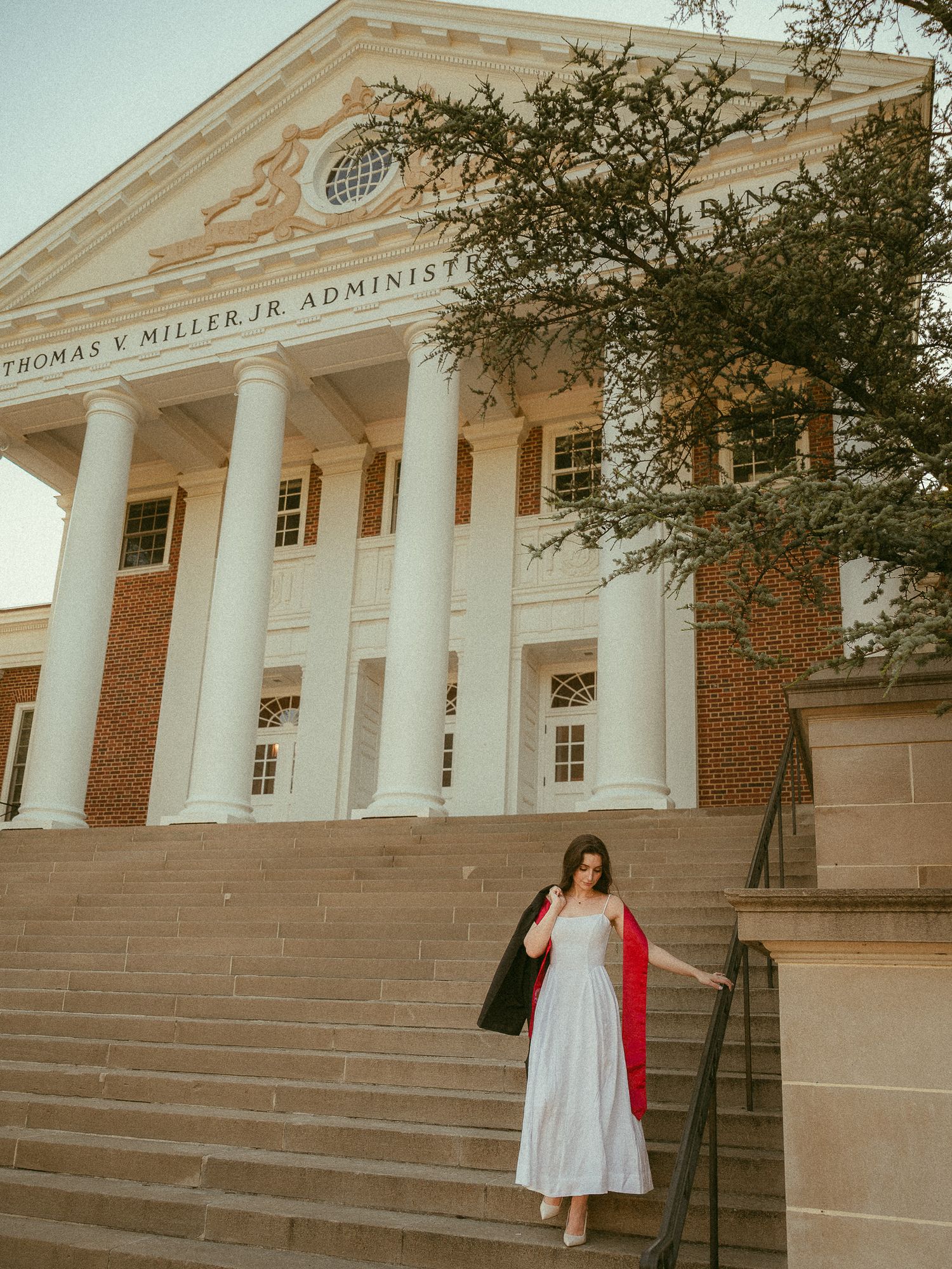 Graduate in white dress descending steps of Thomas V. Miller Jr. Administration building with columns.