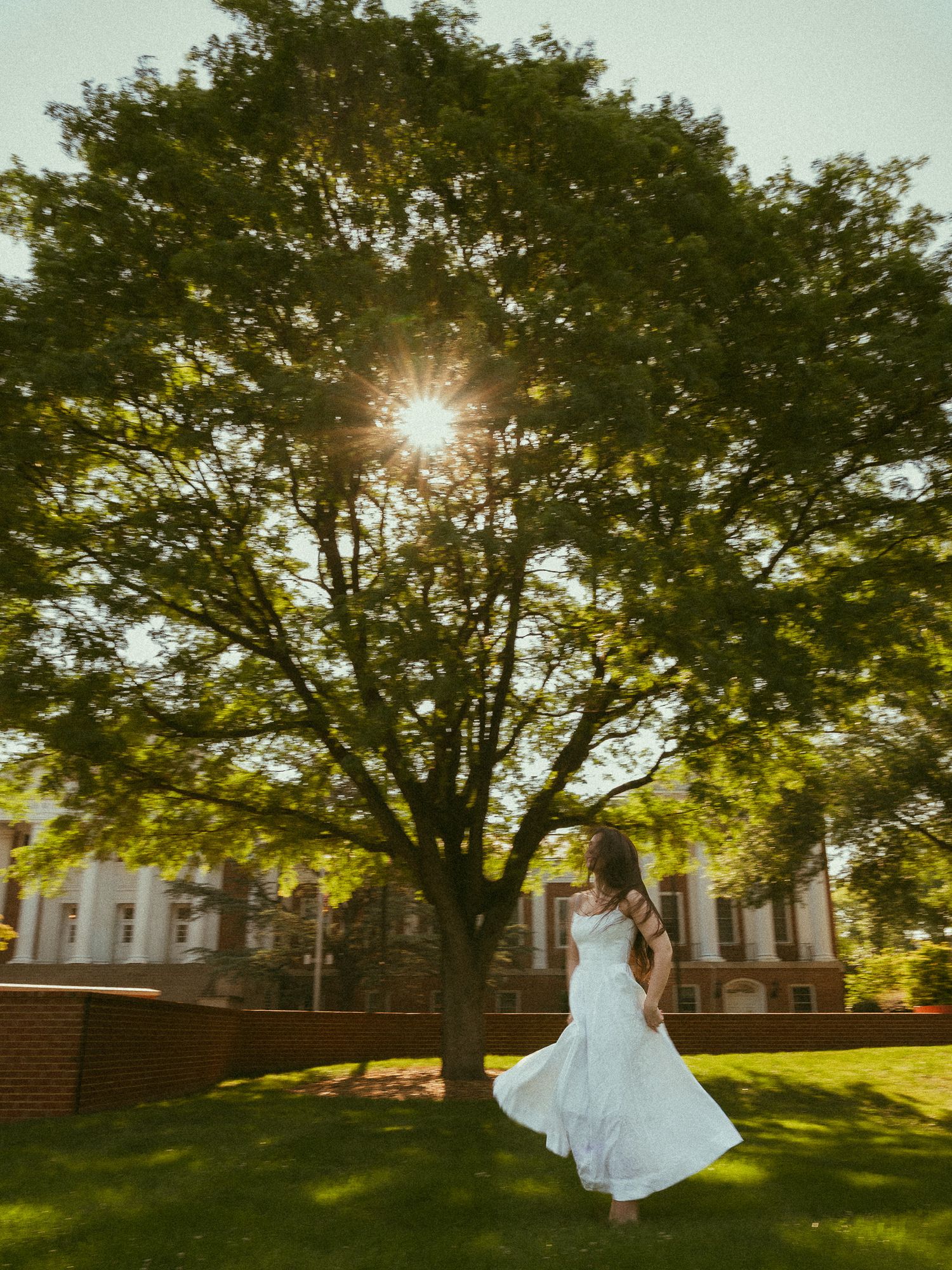 Woman in flowing white dress twirling under a sunburst tree on a college campus green lawn.