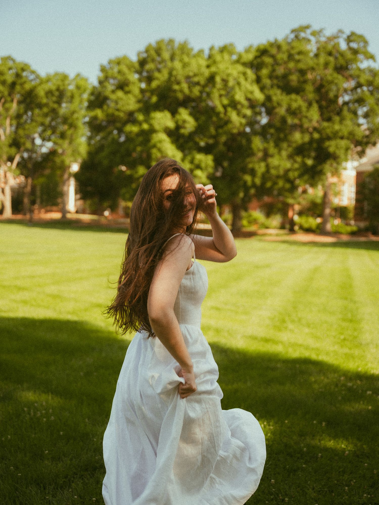Woman in white dress twirling on lush green lawn with trees in background on a sunny day.