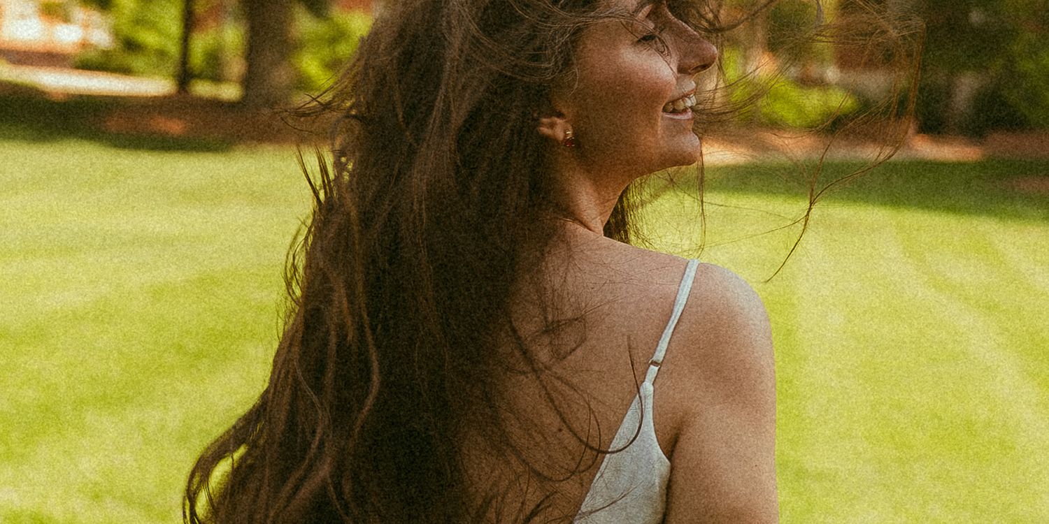 A smiling woman with long flowing dark hair wearing a white sundress, standing outdoors on lush green grass.