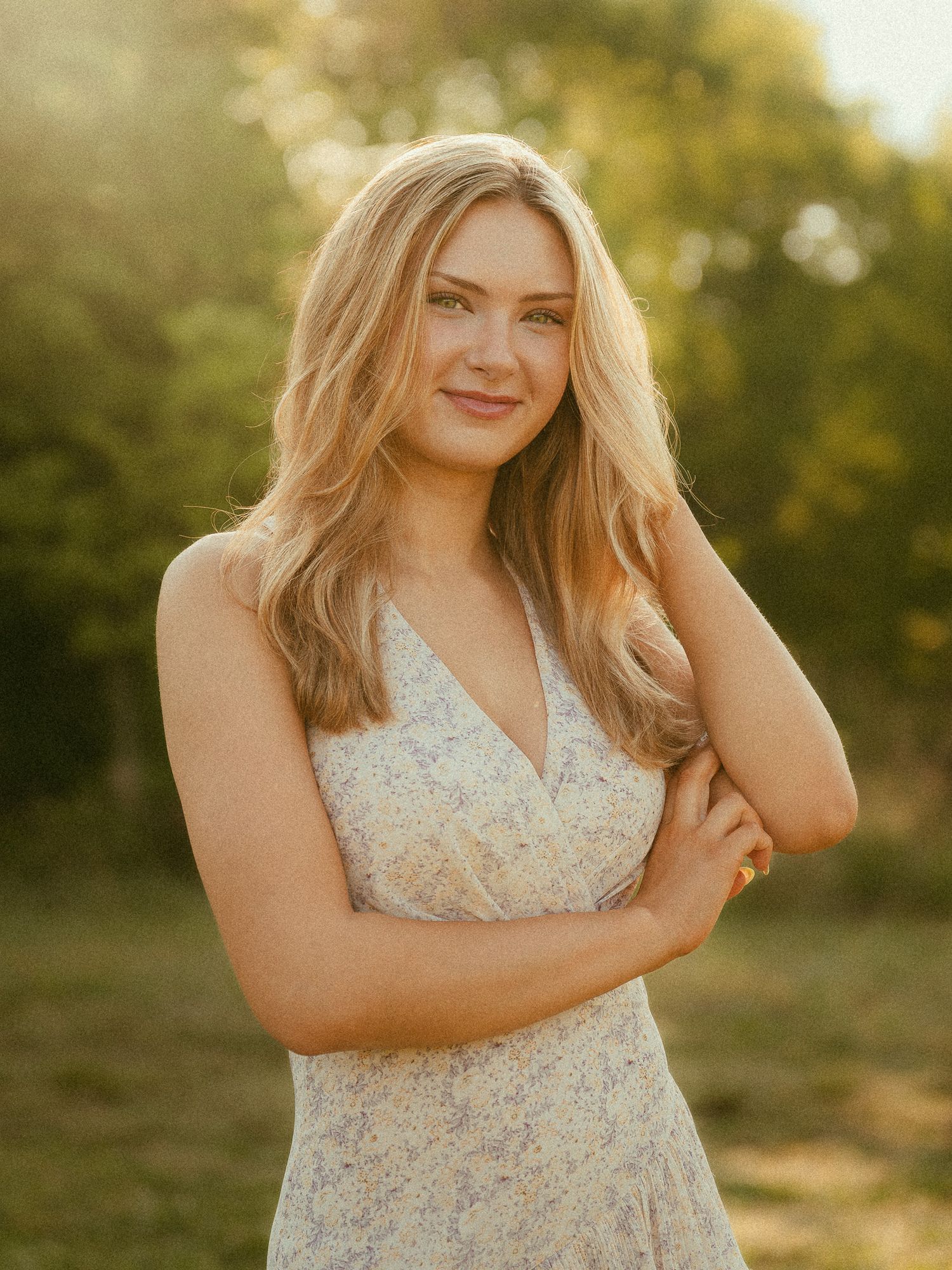 Young woman in floral sundress smiling outdoors in golden hour sunlight with lush green trees in background.