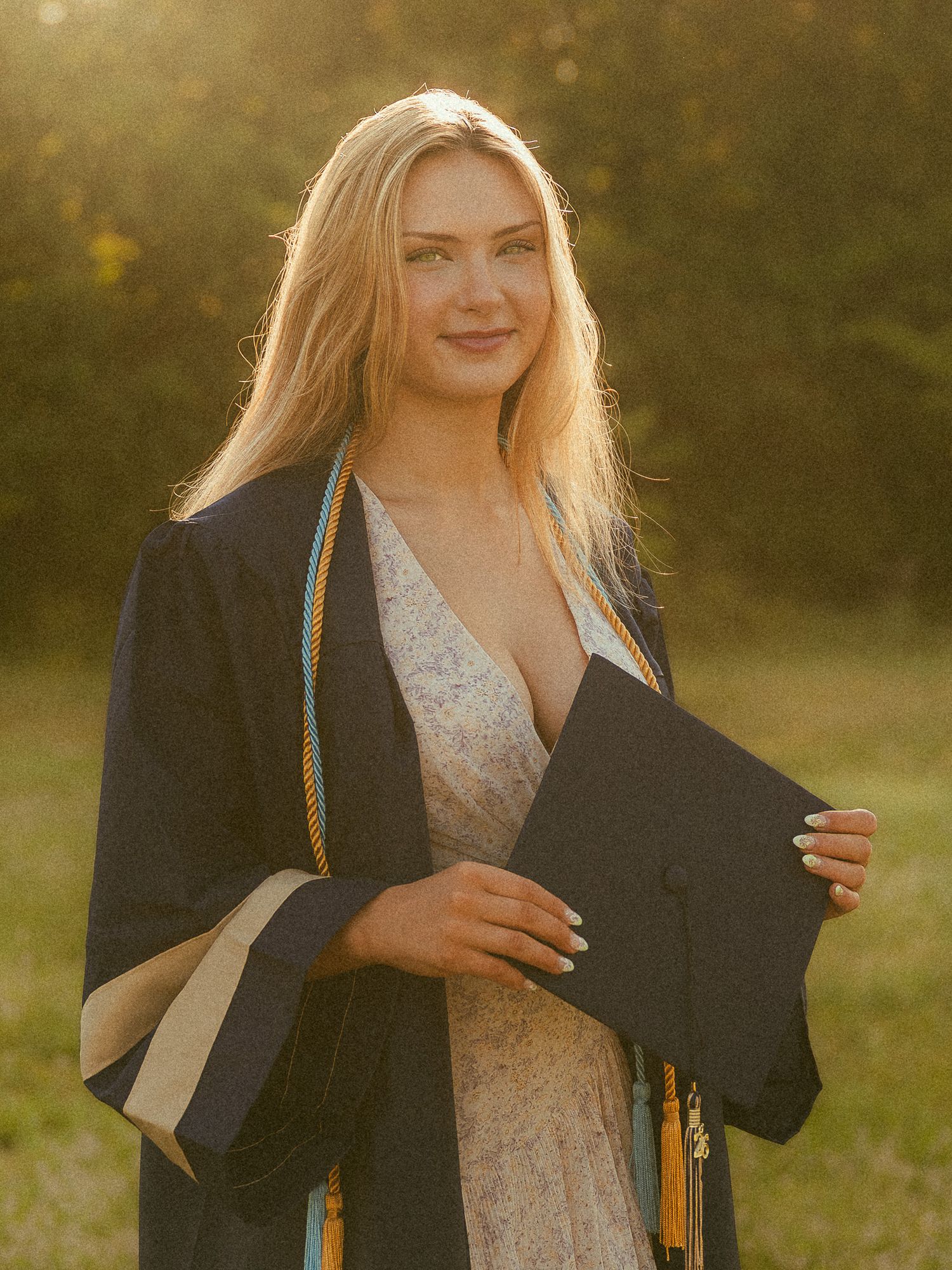 Young woman in graduation gown and cap smiling outdoors in golden sunlight, wearing floral dress with honor cords.