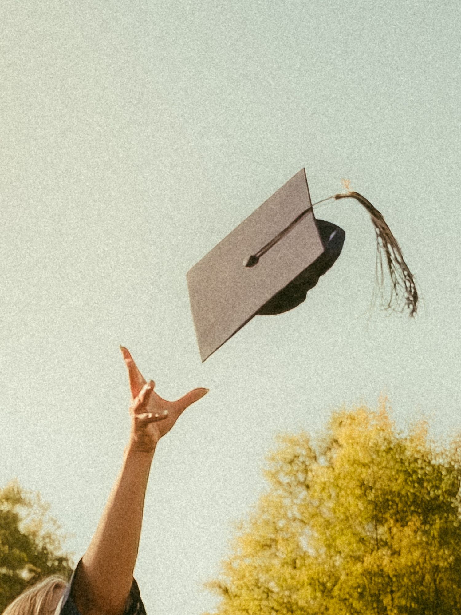 Graduate tossing black mortarboard cap into blue sky with trees in background at graduation ceremony.
