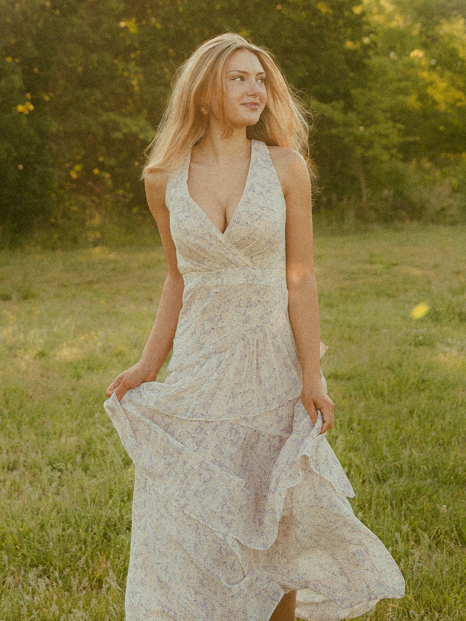 Blonde woman in a floral halter maxi dress standing in a sunlit meadow with green trees in the background.