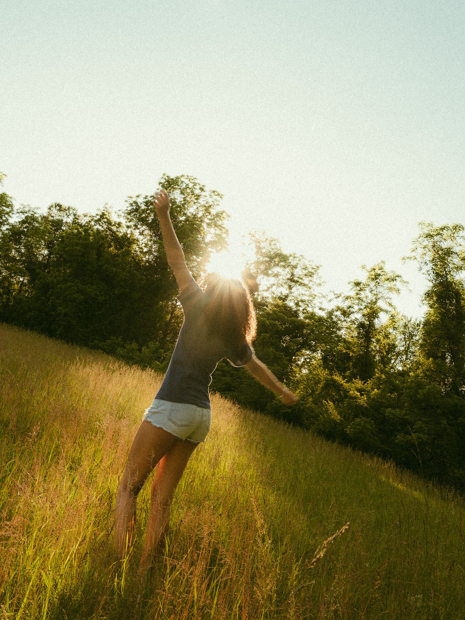 A joyful woman with arms raised stands in a sunlit meadow surrounded by lush green grass and trees.