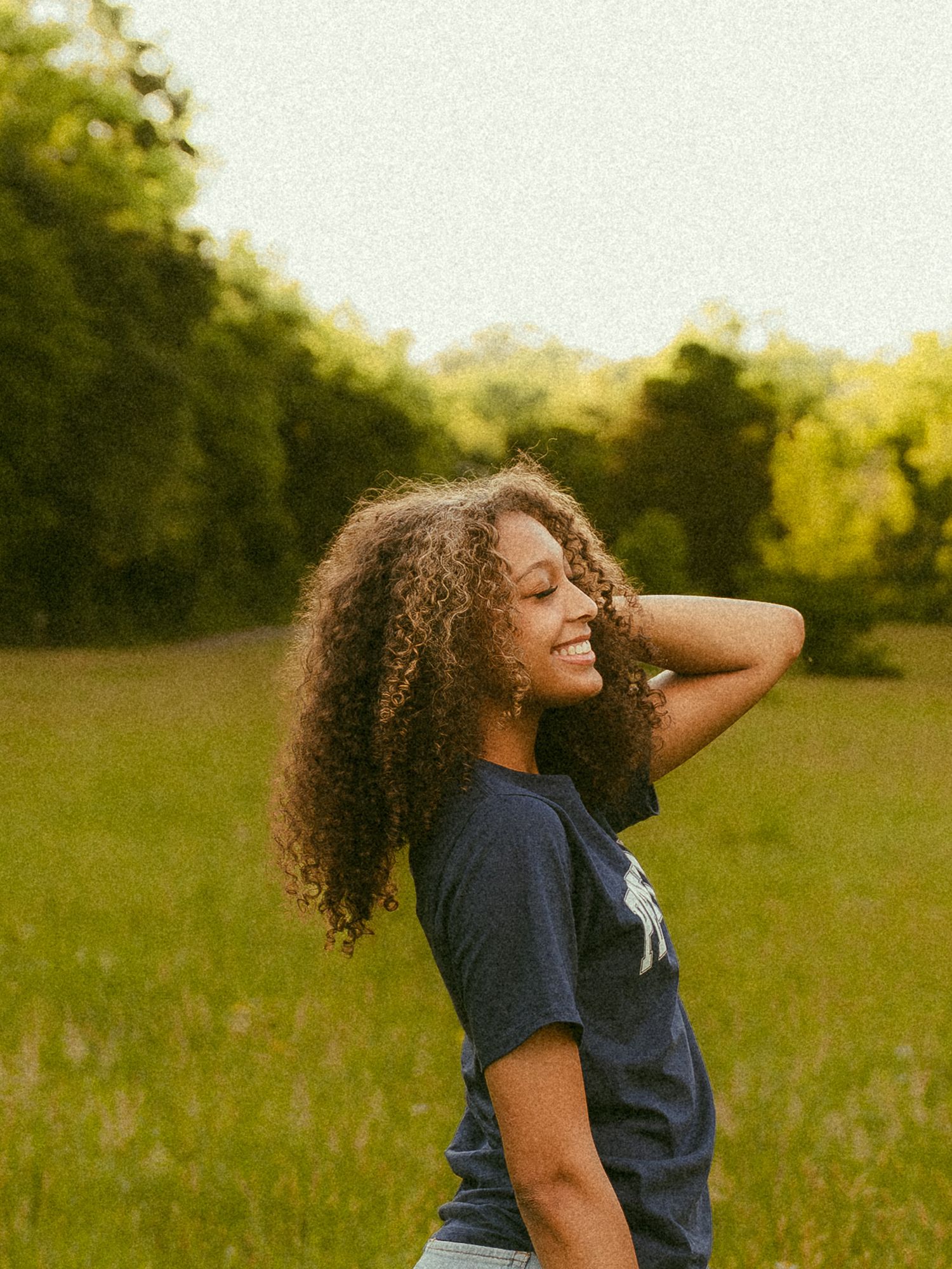 A young woman with curly hair smiles in a green field, wearing a navy t-shirt, with trees in the background.