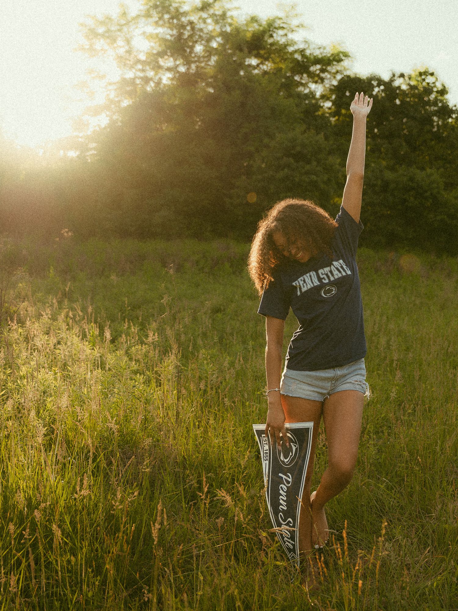 Woman in Penn State shirt and shorts holding a pennant in a sunlit meadow, arm raised joyfully.