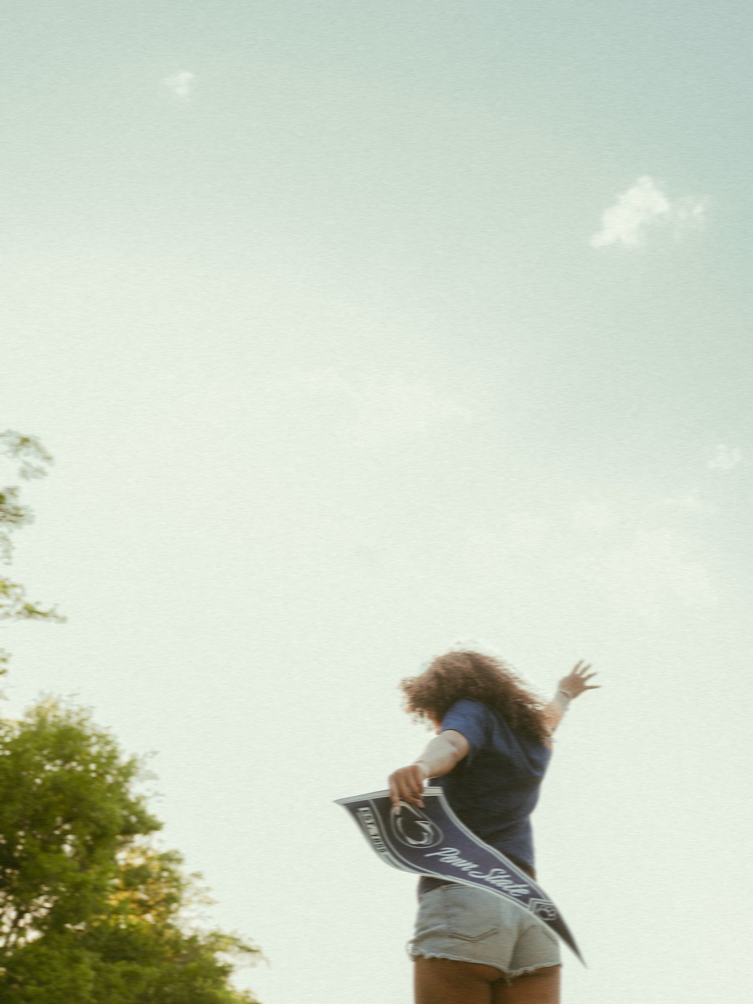 A young woman jumps joyfully outdoors, holding a Penn State pennant against a bright sky with trees nearby.