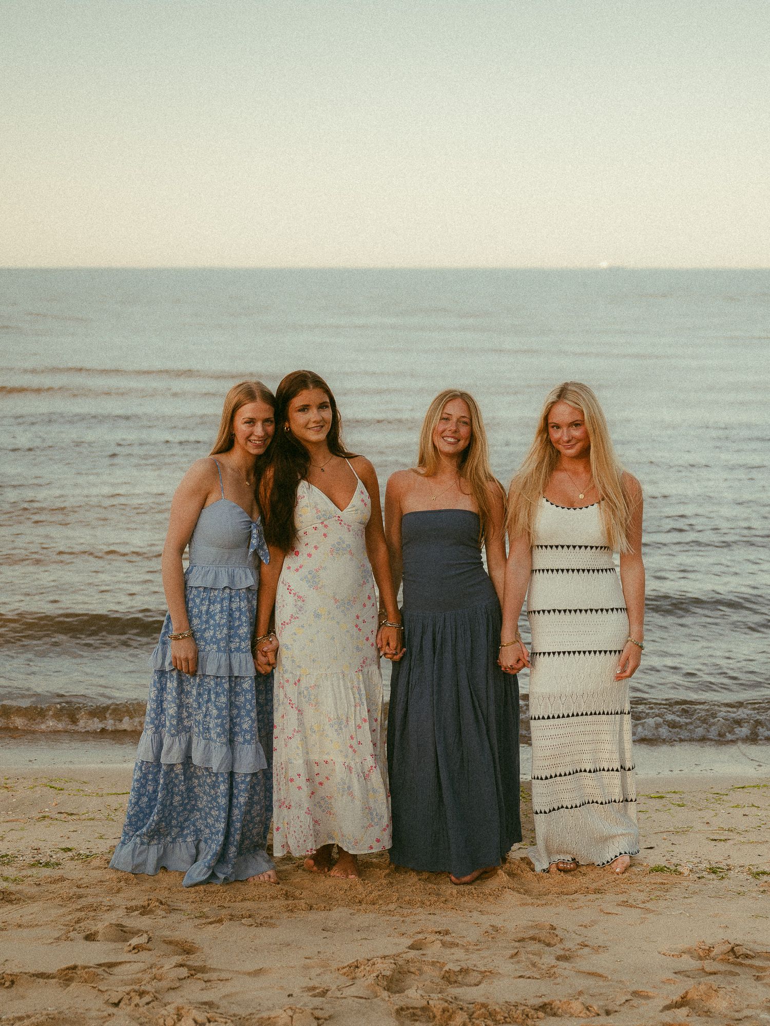 Four young women in maxi dresses pose together on a sandy beach at sunset.