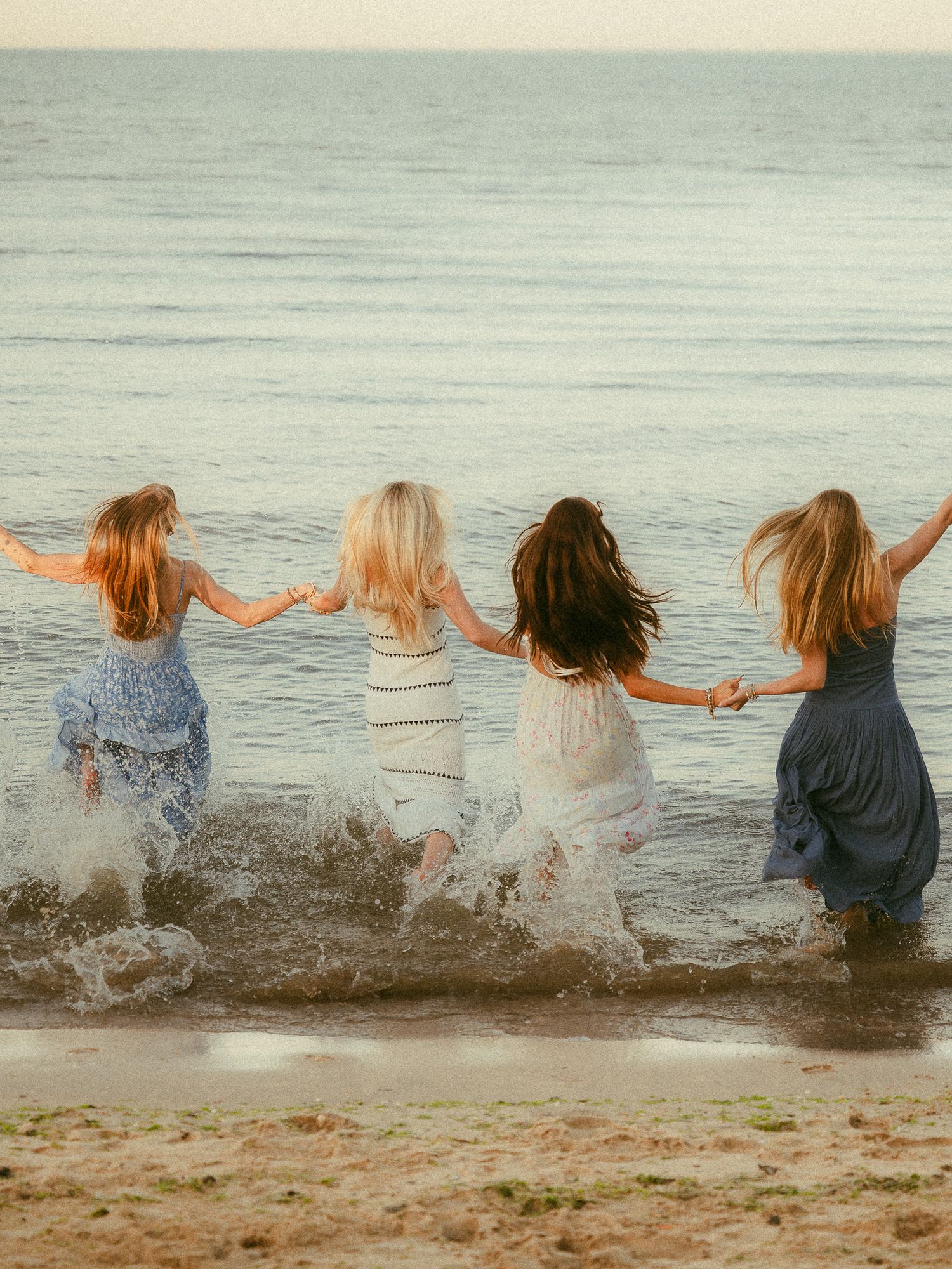 Four girls holding hands running into ocean waves on a sandy beach, wearing flowing dresses, viewed from behind.