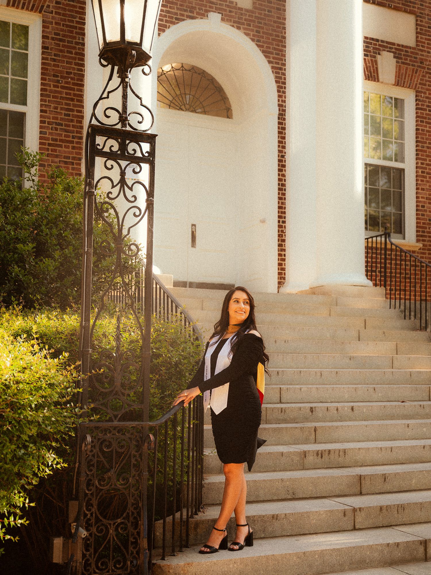 Young woman in black blazer and skirt posing on steps of classic brick building with ornate iron gate.
