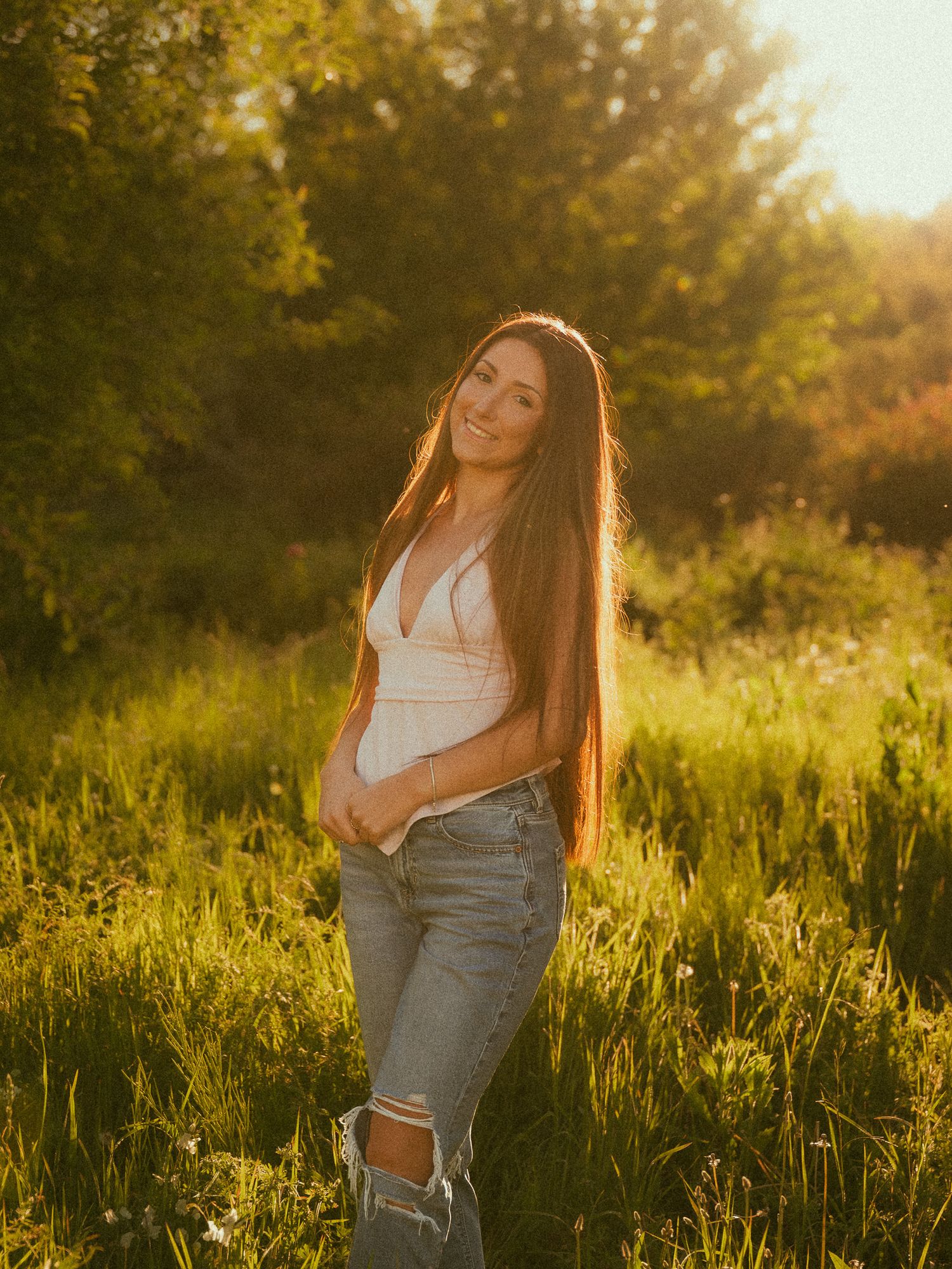 Young woman with long hair in white top and ripped jeans smiling in a sunlit green meadow at golden hour.
